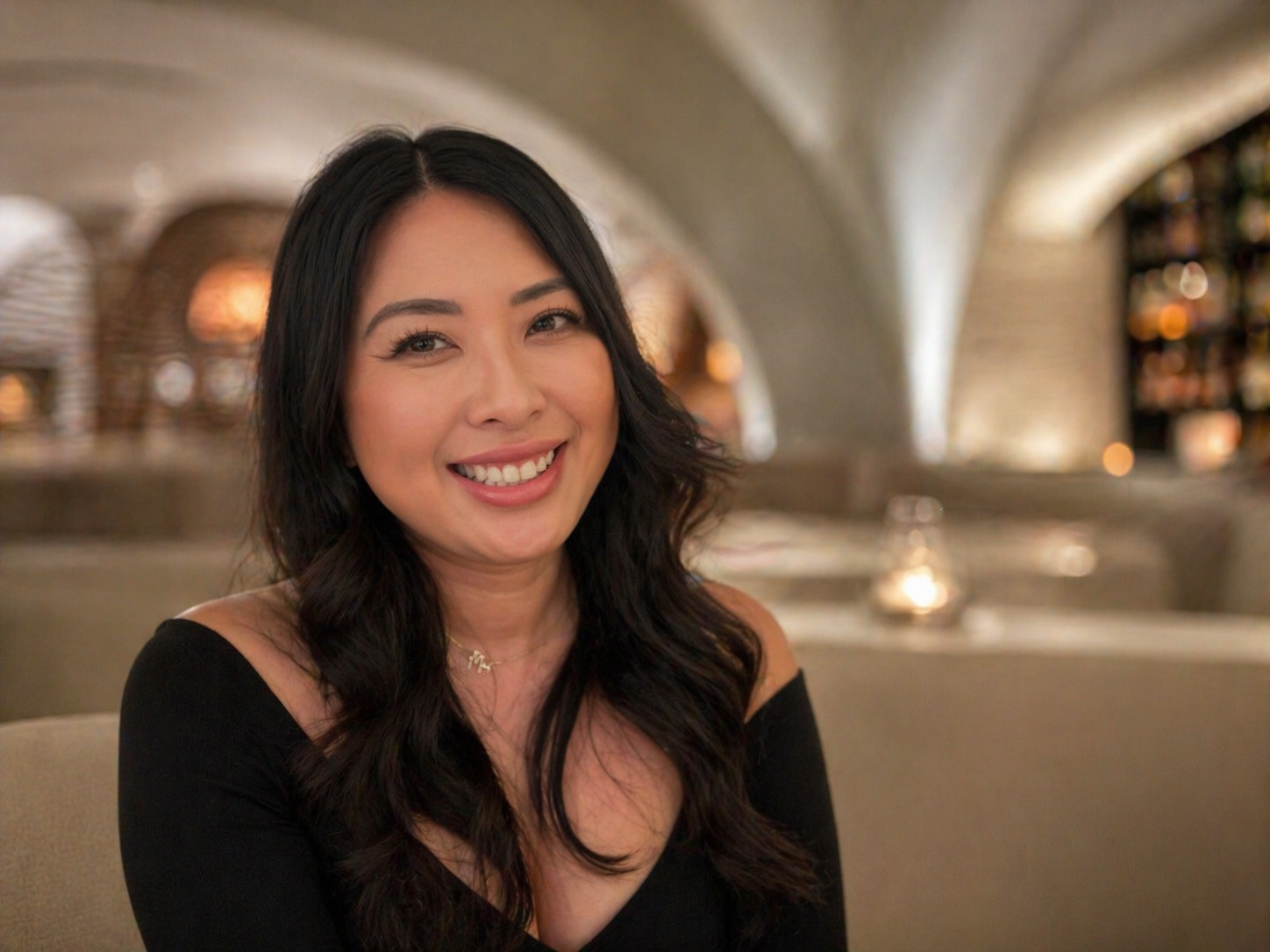 A woman with long dark hair smiling at the camera in a warmly lit indoor setting with arches and blurred background.