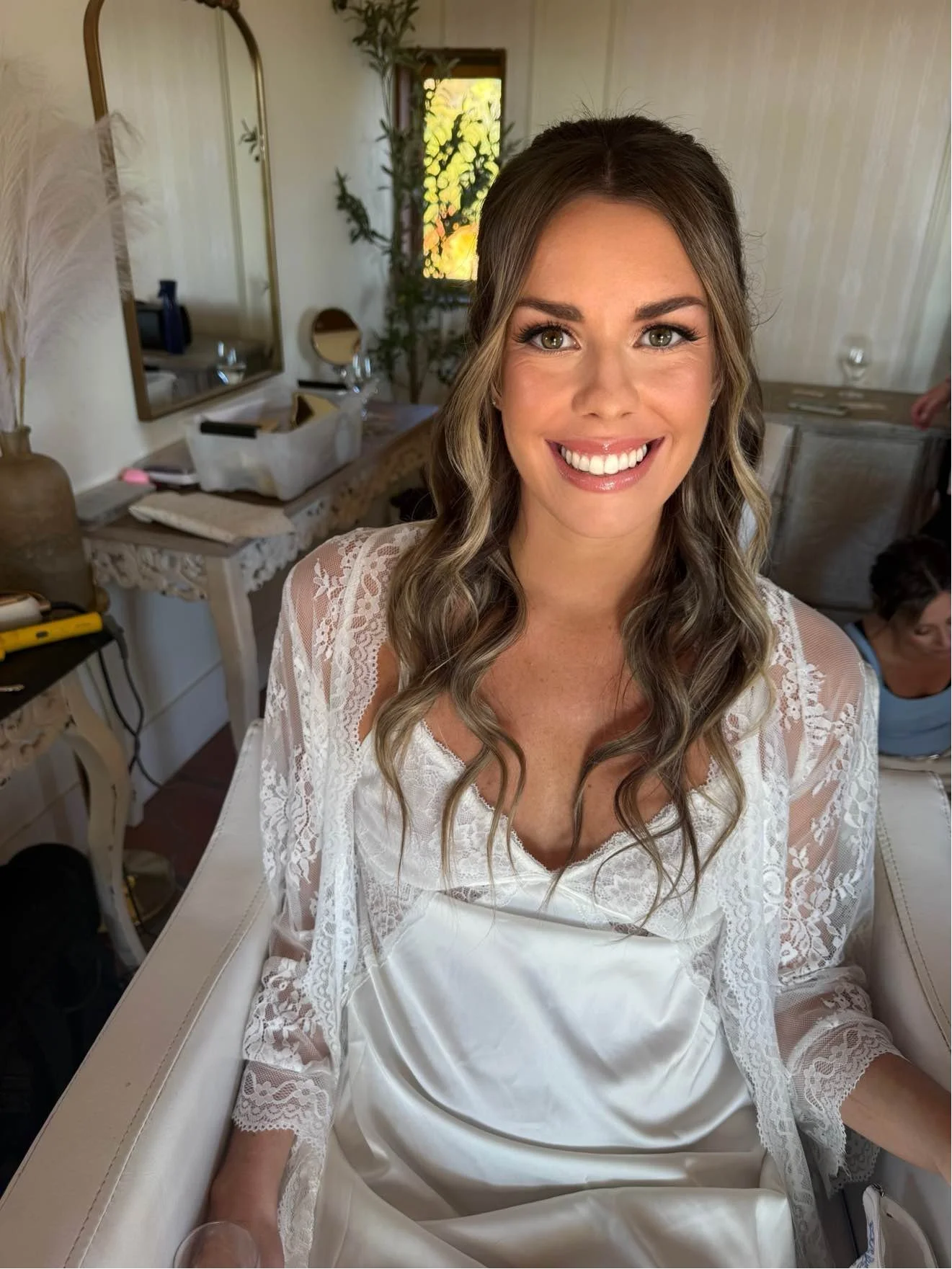 A woman with long wavy brown hair, wearing a white satin and lace dress, smiling with teeth showing, sitting on a cream-colored chair inside a room with wooden furniture and a mirror, with another person partially visible in the background.