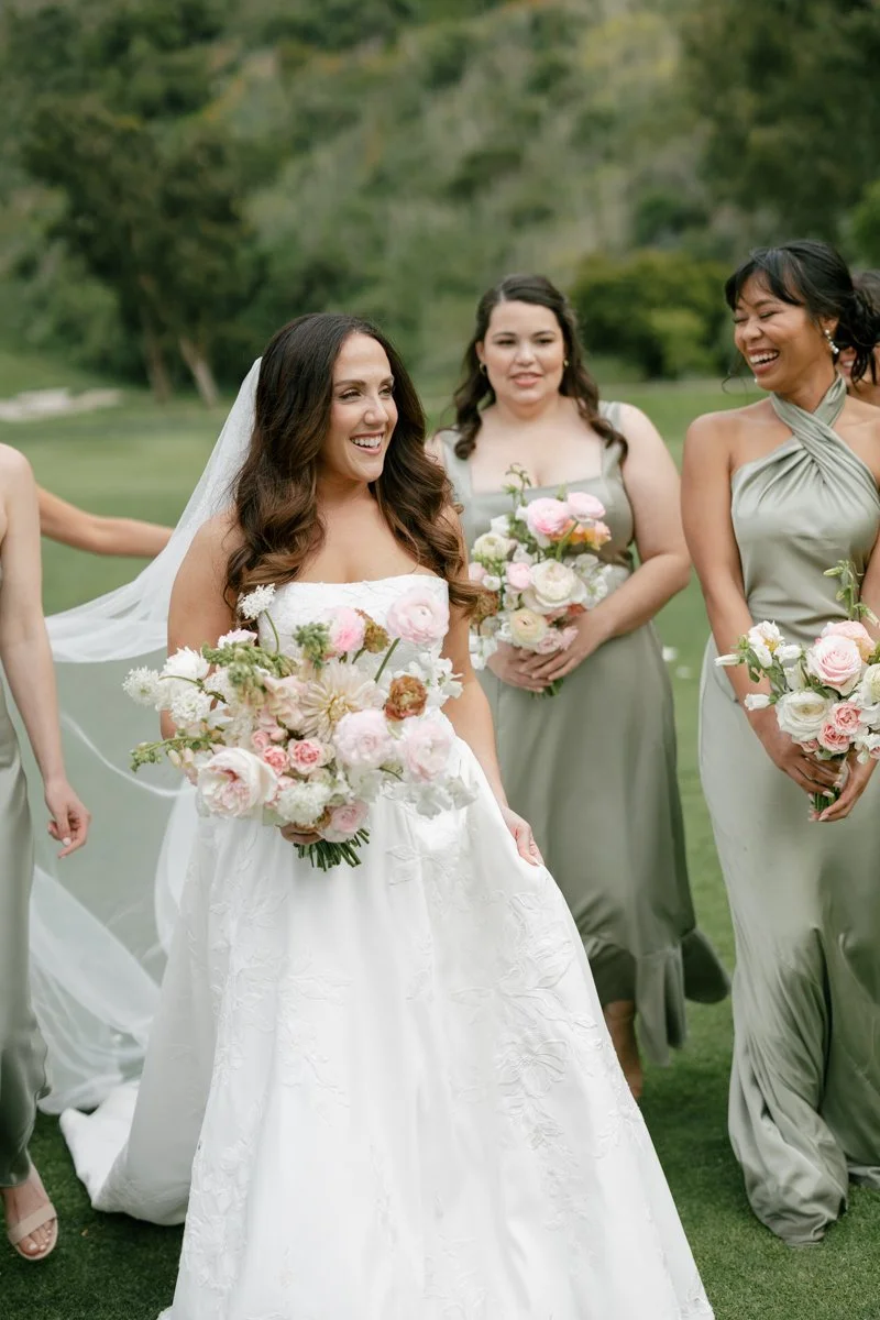 A bride in a white wedding dress holding a bouquet of pink and white flowers, smiling, with bridesmaids in gray dresses holding similar bouquets, outdoors on a grassy field with trees in the background.