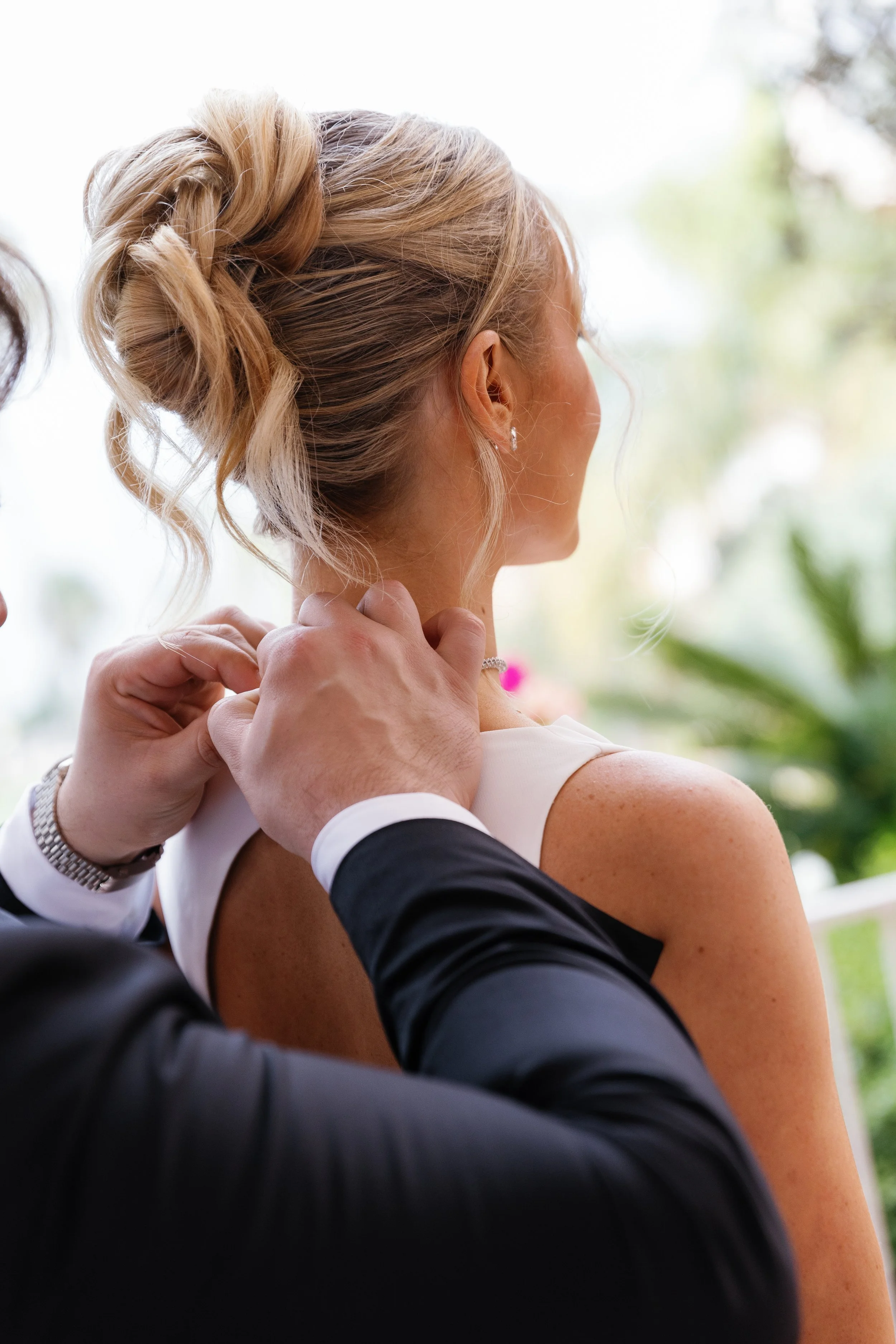 A woman with blonde hair styled in an elegant updo, wearing earrings, is being helped by a person in a suit to adjust her necklace. The scene appears to be at a formal event, possibly a wedding or celebration.