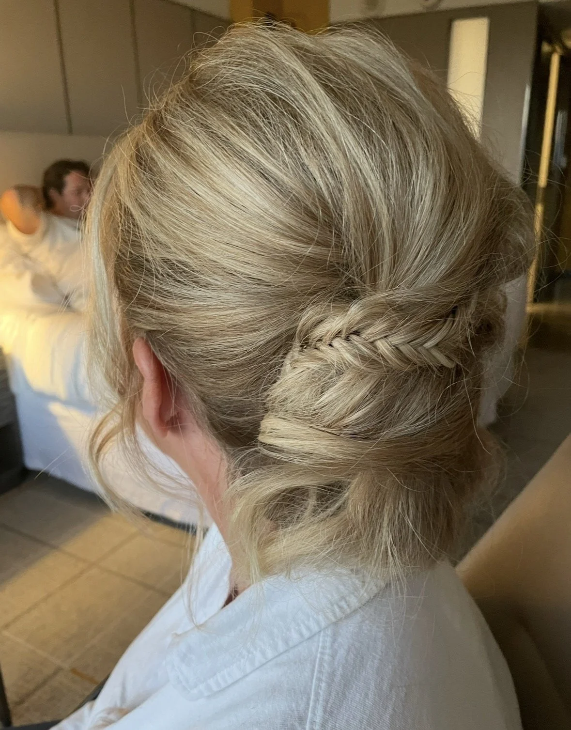 Back of a woman with blonde hair styled in a twisted updo, sitting in a room with someone in the background.