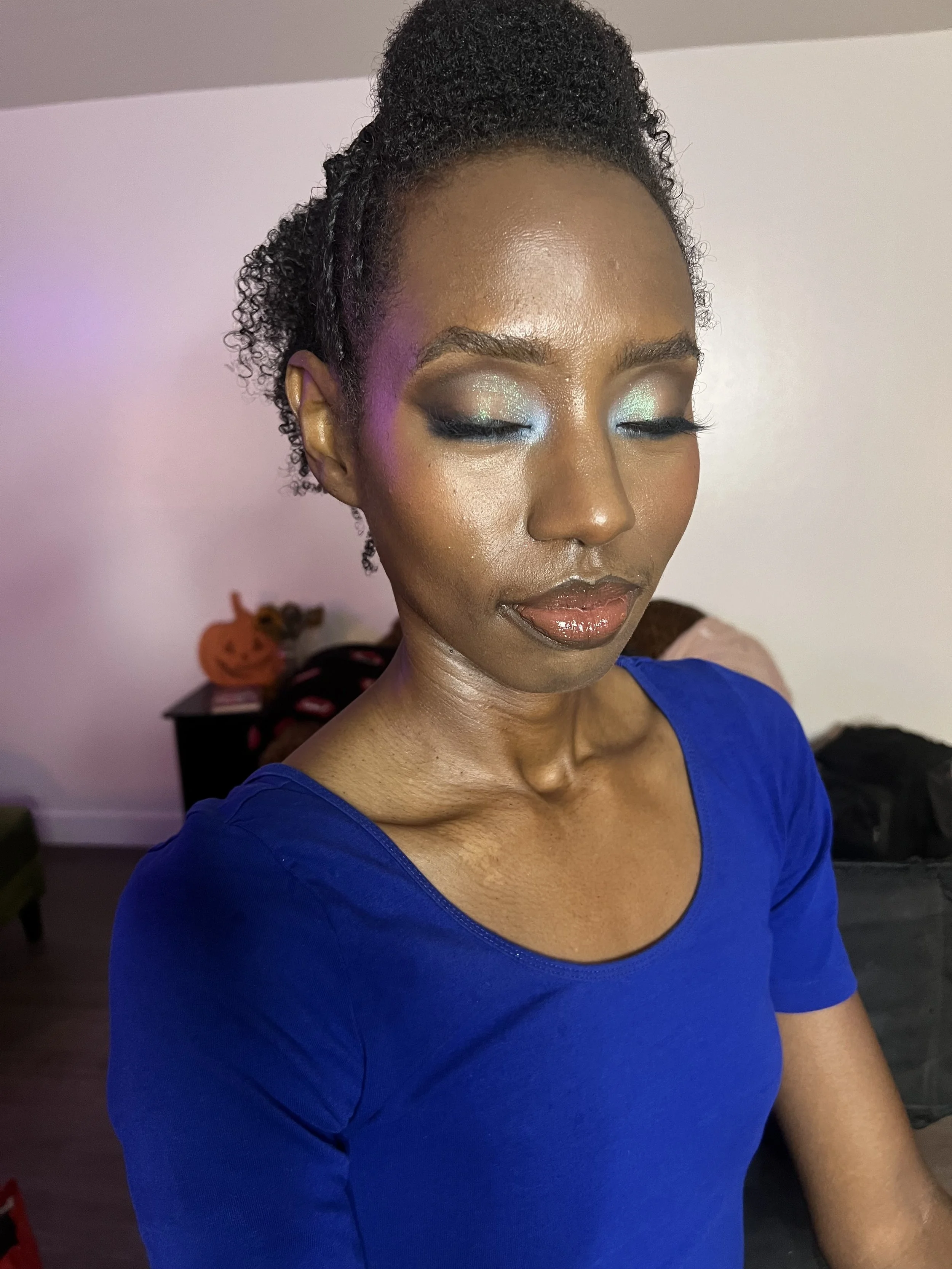 A woman with closed eyes and curly hair styled in an updo, wearing makeup with shimmering eyeshadow and gloss, dressed in a blue top, standing indoors with a pumpkin decoration in the background.