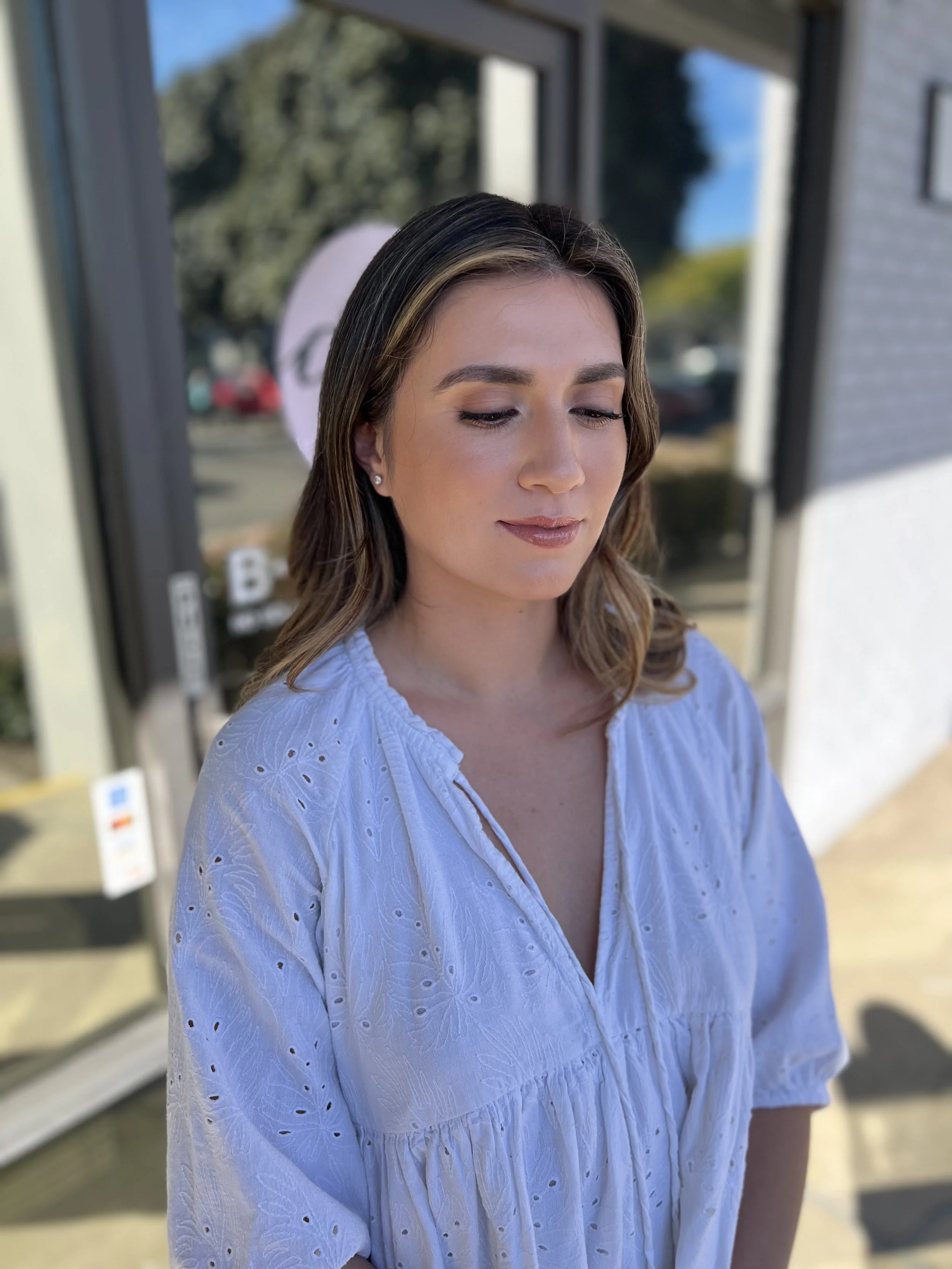 A young woman with long wavy hair, wearing a white embroidered blouse, stands outdoors near a storefront, looking down with a serene expression.