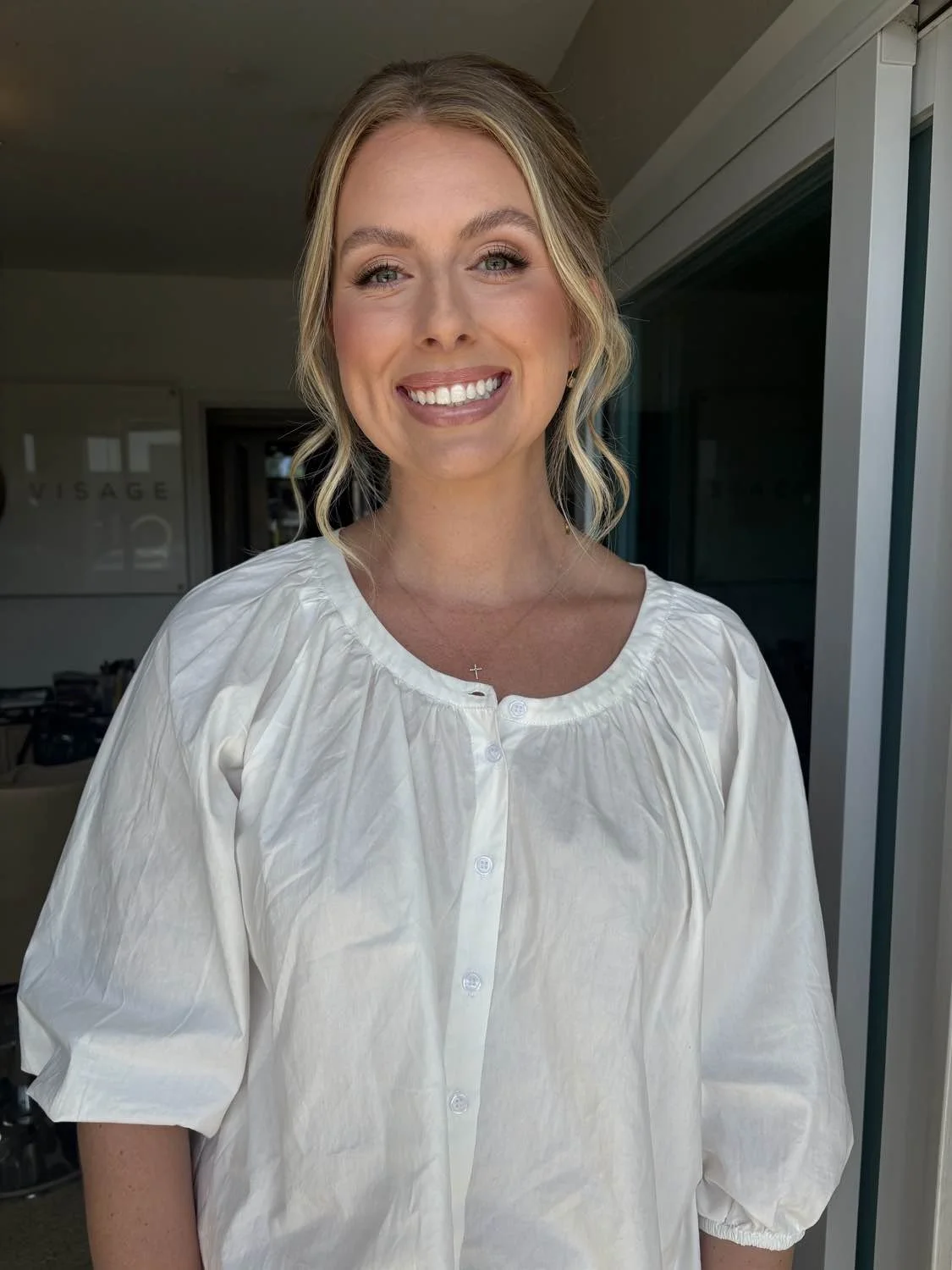 A woman with blonde, shoulder-length wavy hair wearing a white button-up blouse standing indoors near a sliding glass door, smiling at the camera.