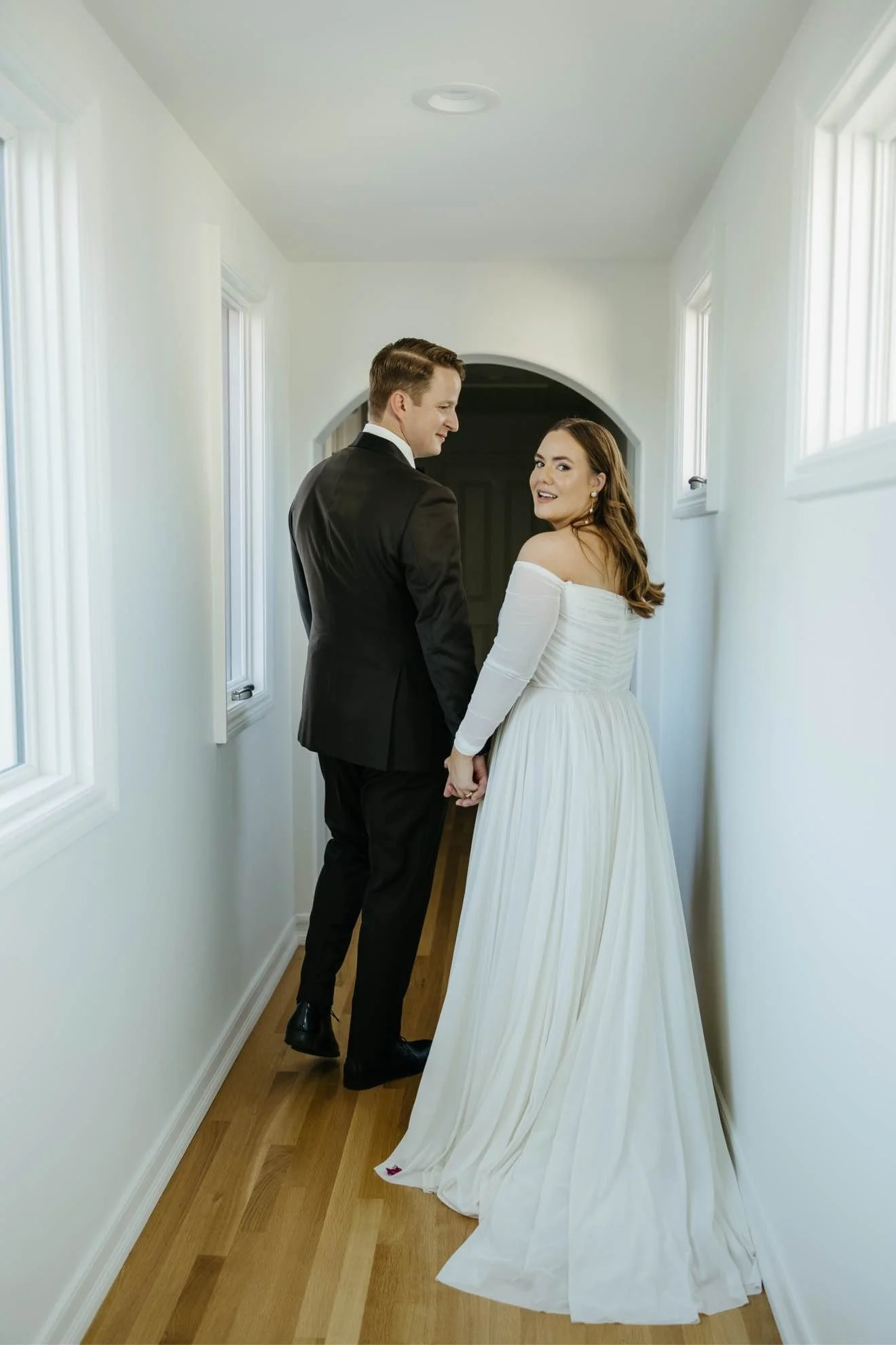 A couple dressed in wedding attire holding hands and walking down a narrow hallway with white walls and windows.
