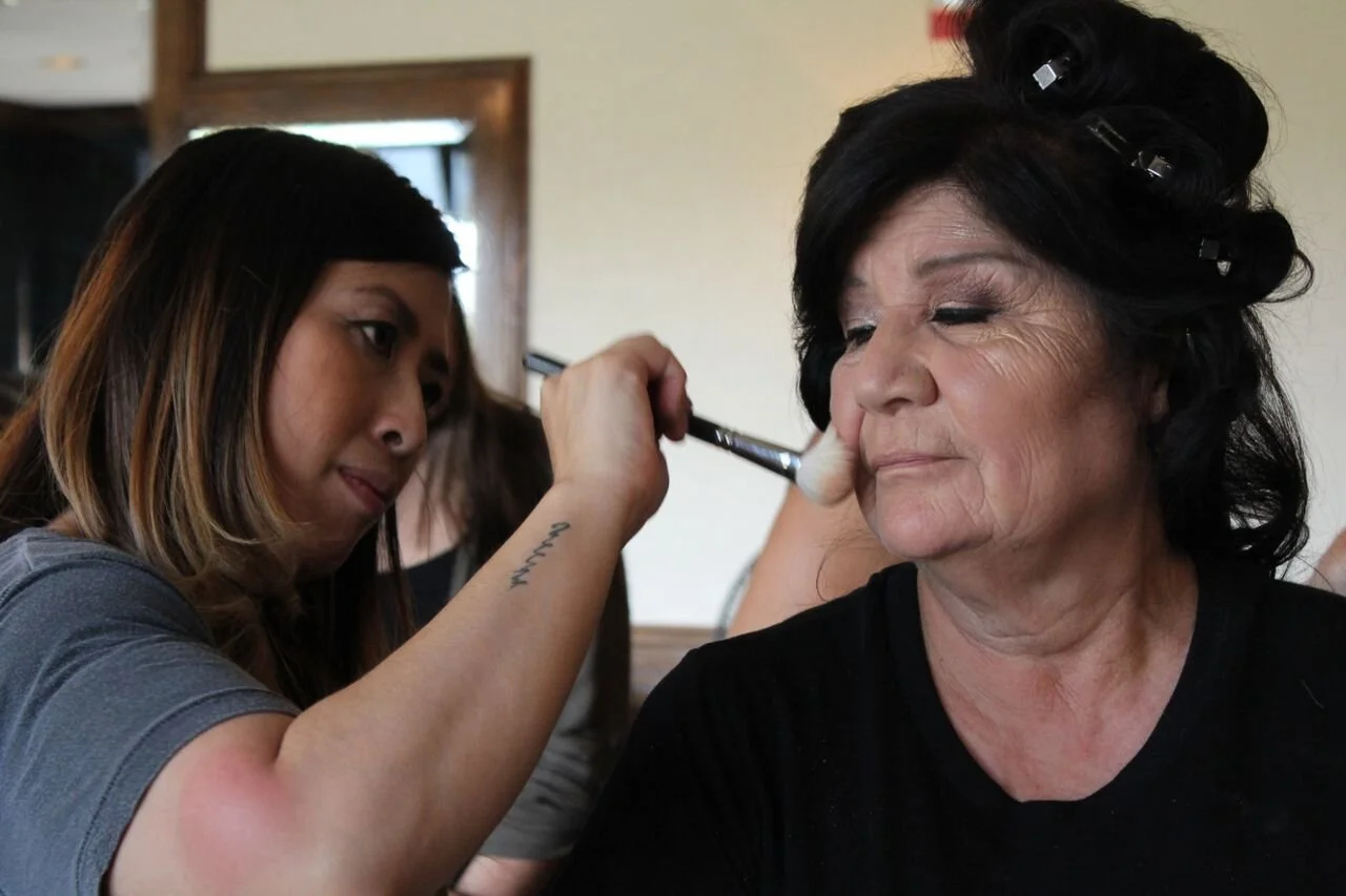 A woman applying makeup to an elderly woman with rollers in her hair inside a room.