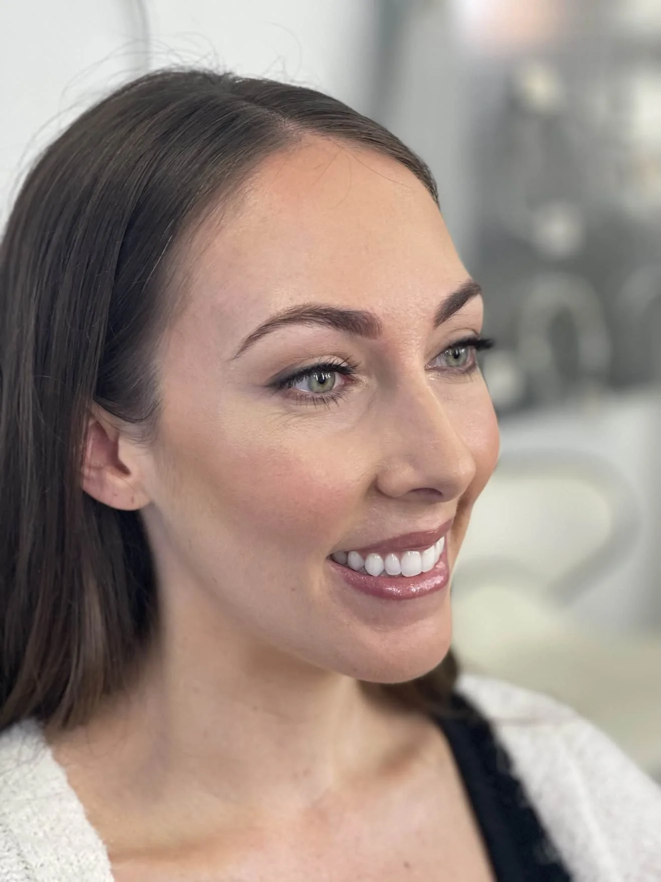 A smiling woman with long dark hair and blue eyes, wearing makeup, in a bright, salon or studio setting.