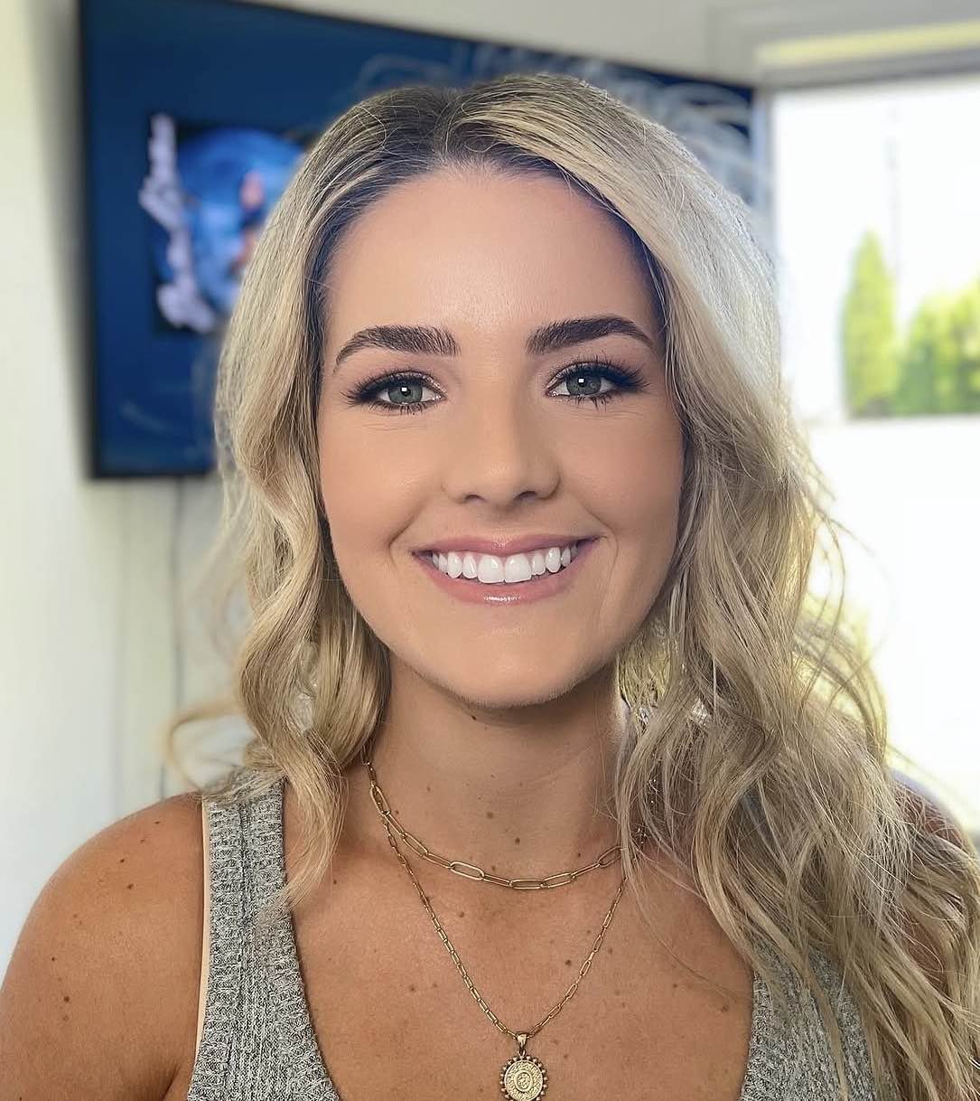 A young woman with blonde wavy hair smiling, wearing layered necklaces, a gray sleeveless top, and makeup, standing in front of a TV and window with green trees outside.
