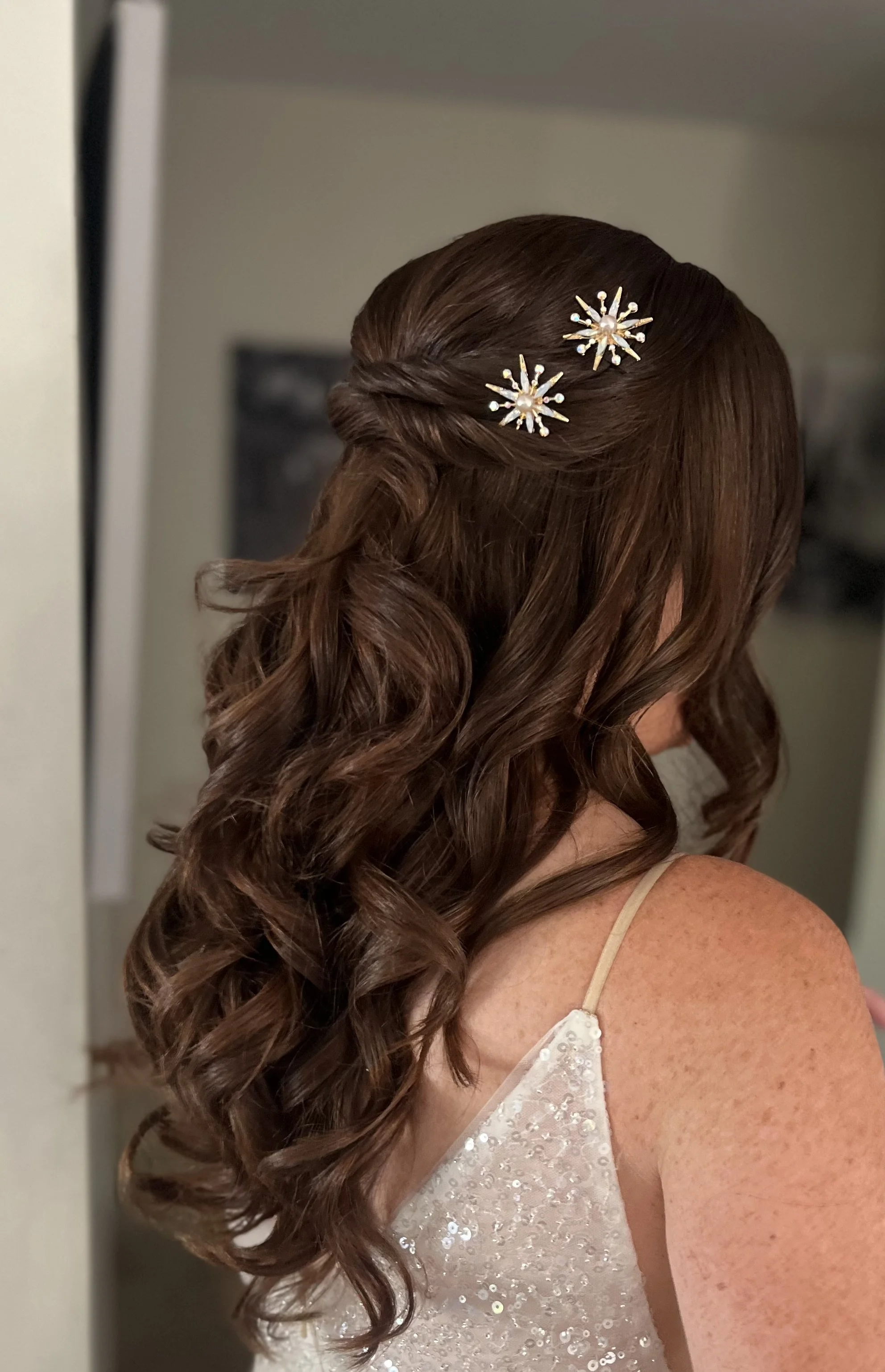 Close-up of a woman with curly brown hair styled with two decorative pearl and gold starburst hairpins, wearing a white, sequined dress with thin straps.
