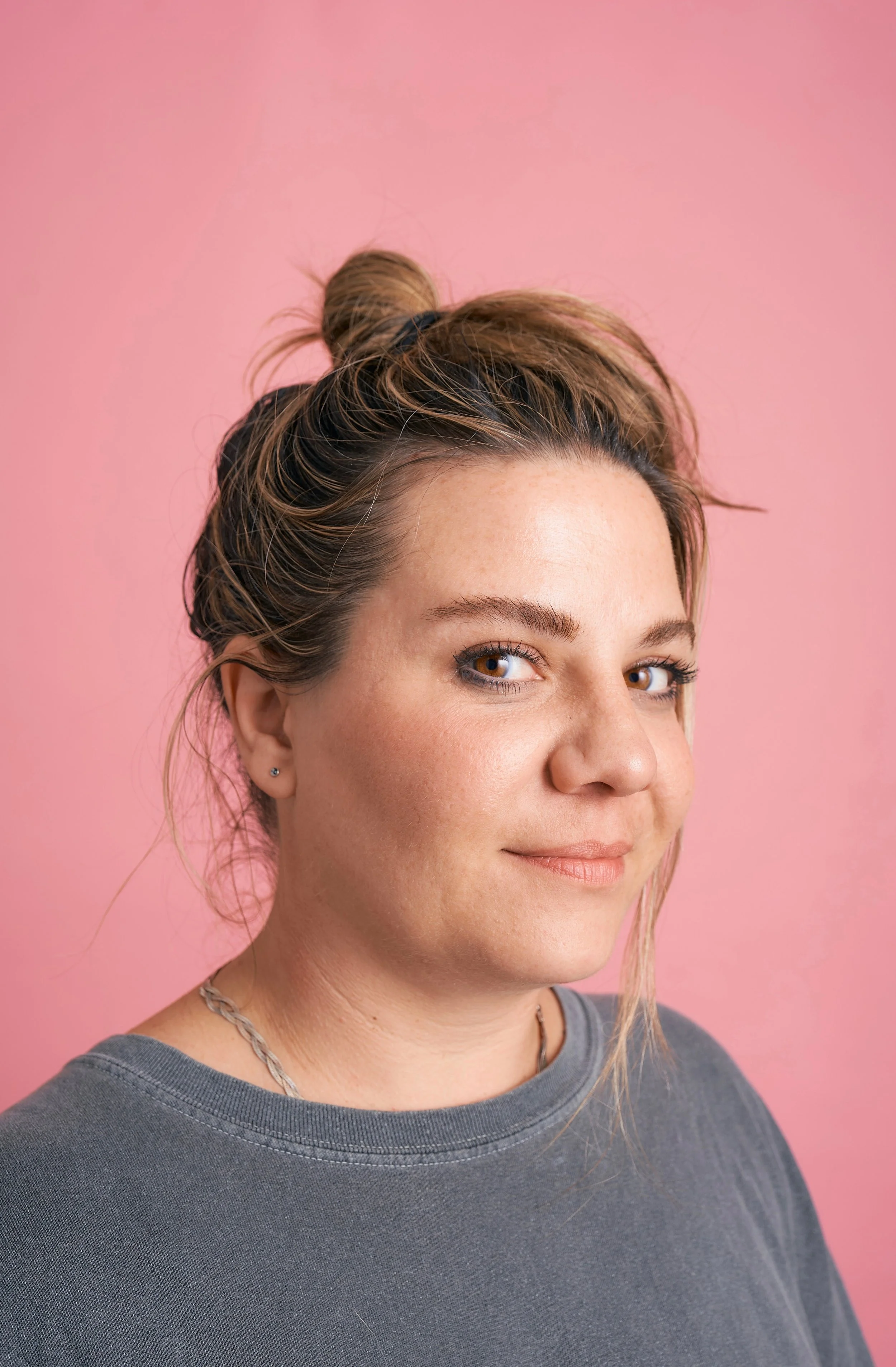 Close-up of a woman with brown hair in a bun, smiling slightly, against a pink background, wearing a gray shirt and a silver chain necklace.