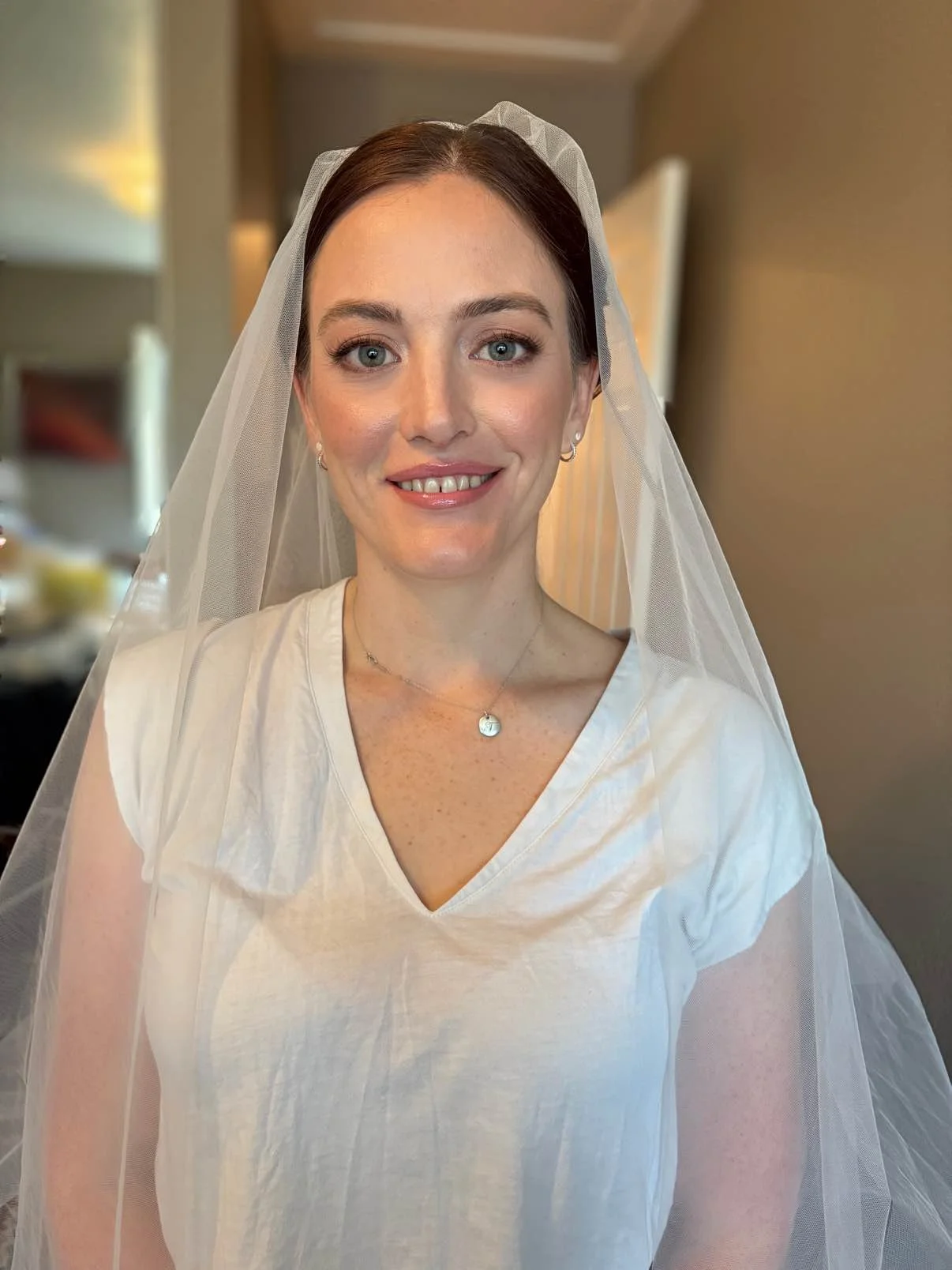 A woman with blue eyes and brown hair wearing a white t-shirt, a necklace, earrings, and a veil, smiling at the camera inside a room.
