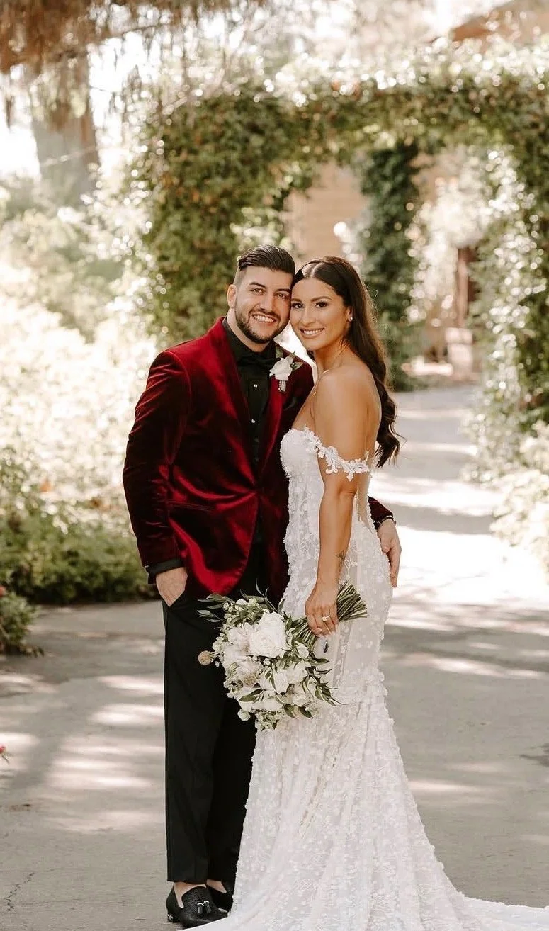 A bride and groom smiling and posing together outdoors during their wedding, with greenery and an arch in the background. The bride is wearing a white lace wedding dress and holding a bouquet of flowers, while the groom is dressed in a dark suit with