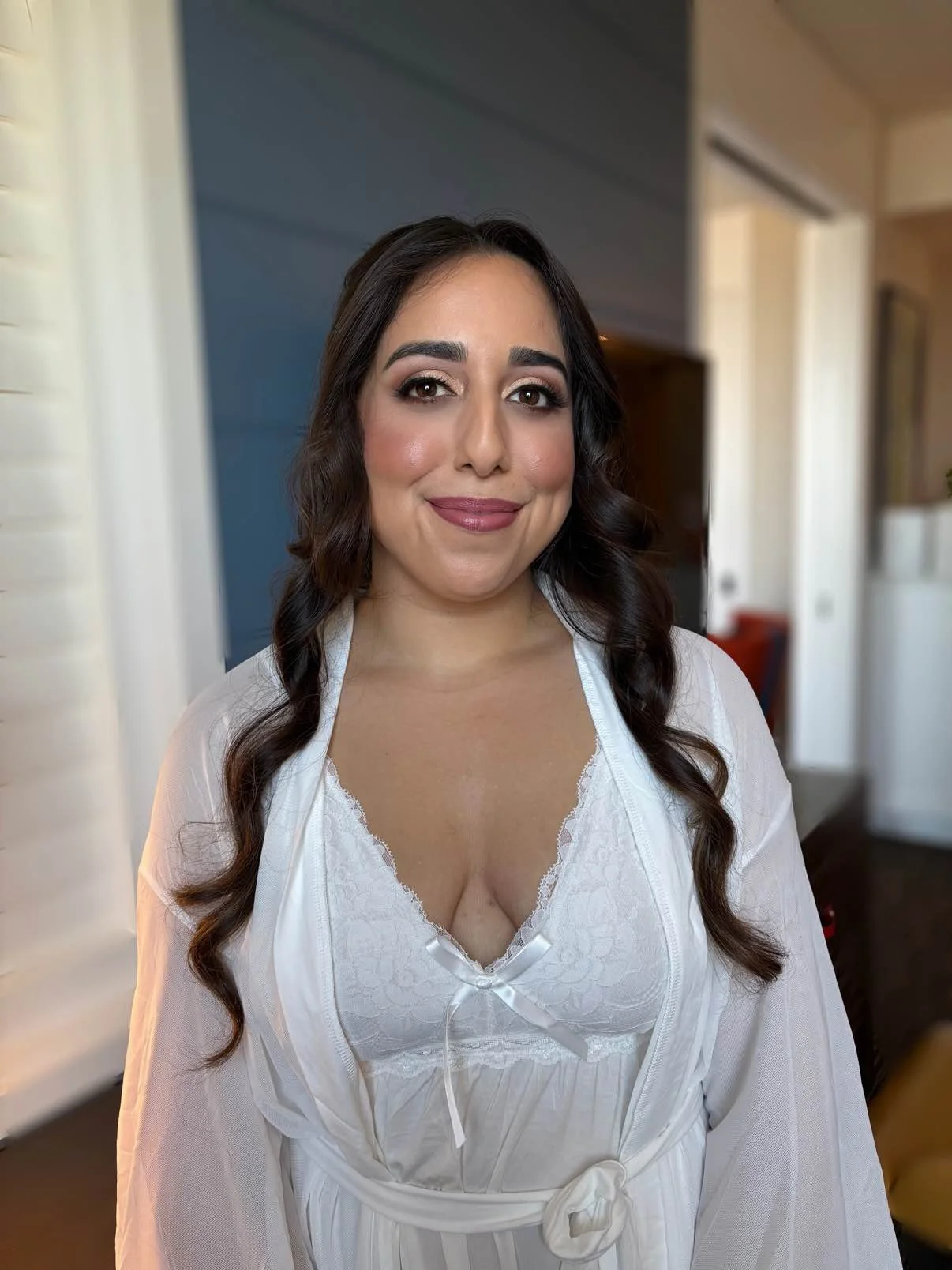 A woman with long dark wavy hair and makeup, wearing a white lace-trimmed top and a sheer white robe, standing indoors with a blurred background.