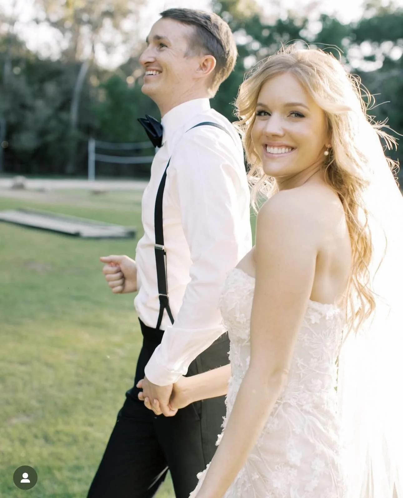 A smiling bride and groom holding hands outdoors during daylight, with the bride in a lace wedding dress and the groom in a white shirt with suspenders and a bow tie, standing on a grassy area.