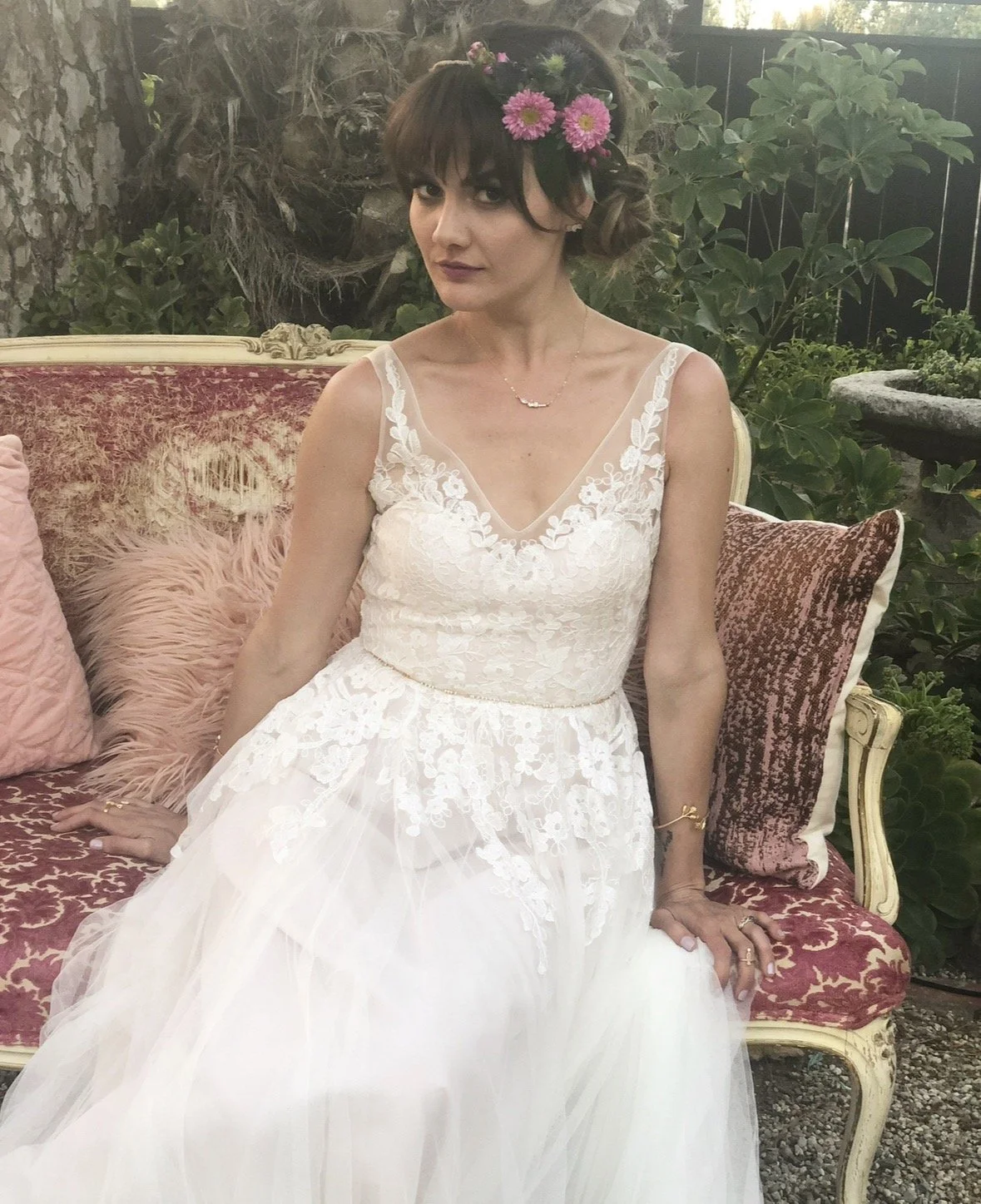 Woman in a white wedding dress sitting on a vintage-style pink and gold couch with decorative pillows, outdoors with greenery and trees in the background, wearing a floral headpiece and jewelry.