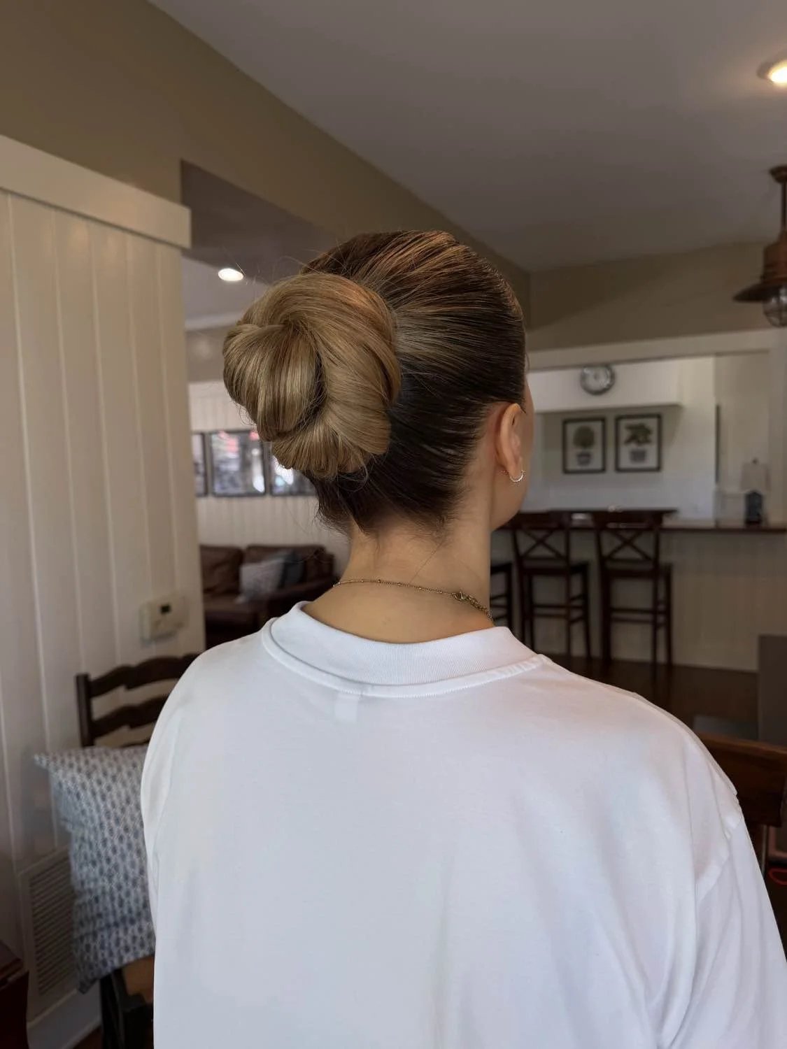 Rear view of a woman with a neatly styled bun hairstyle, wearing a white shirt and jewelry, inside a cozy room with dining and living areas visible.