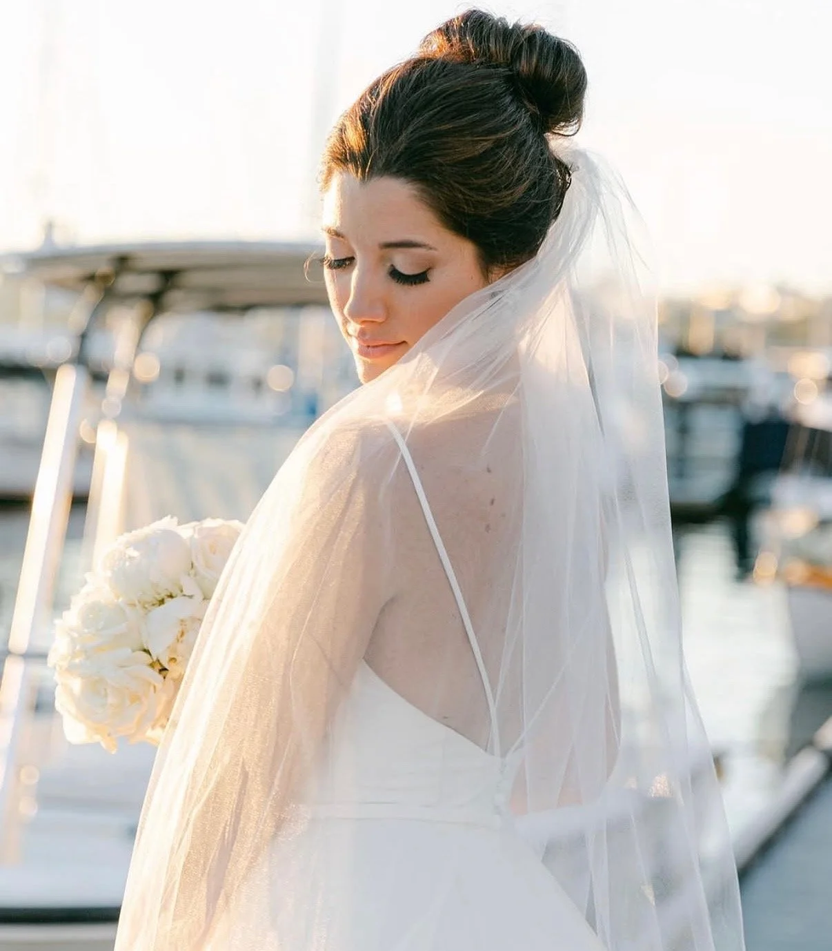 A bride in a white wedding dress with a veil, holding a bouquet of white roses, standing near boats at a marina during sunset.