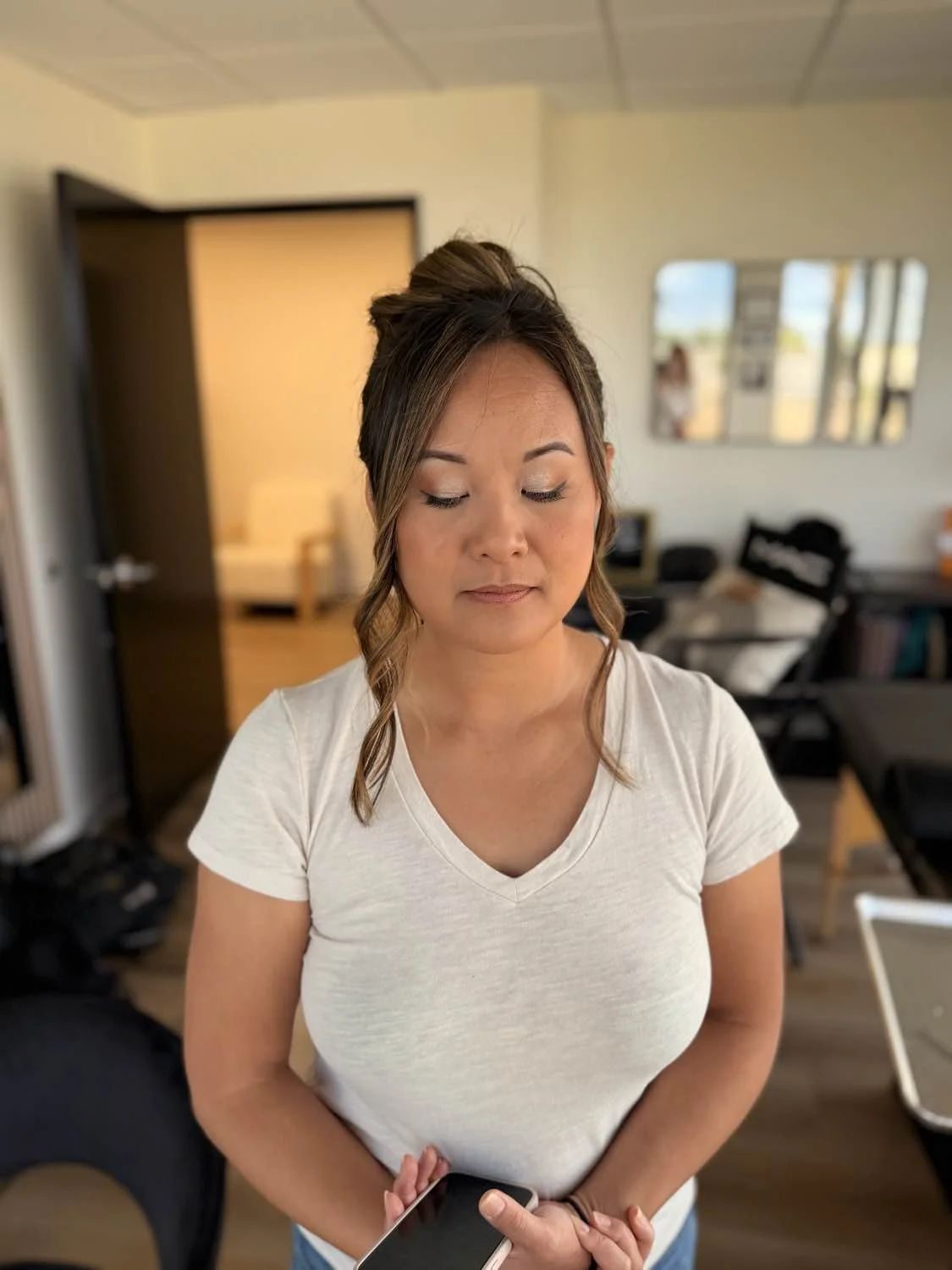 A woman with medium-length hair styled in waves and a half-up bun, wearing a white V-neck T-shirt, standing in a room with a relaxed expression, holding a smartphone in her hands, with a blurred background of a living room and furniture.
