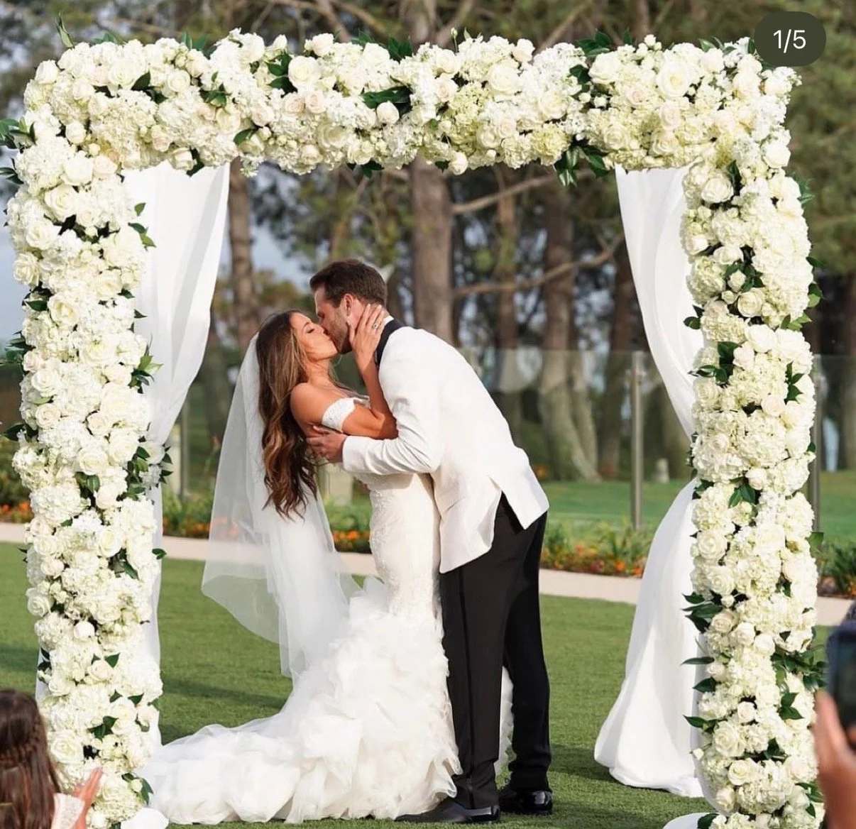 Couple kissing under a white floral wedding arch outdoors.
