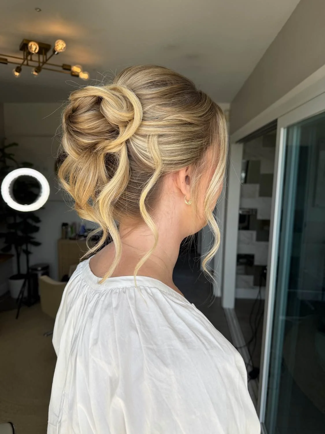 A woman with blonde hair styled in an elegant updo with loose curls, wearing a white blouse and small hoop earrings, standing in a modern indoor space with a ring light and a sliding glass door.