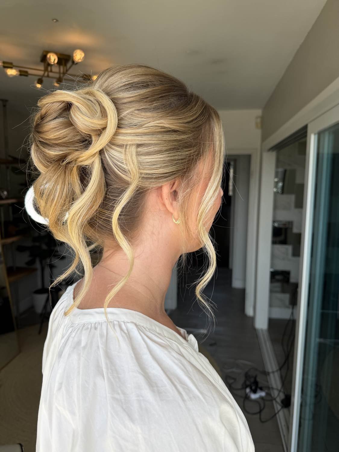 A woman with blonde hair styled in an elegant updo featuring loose curls, viewed from the side in an indoor setting.