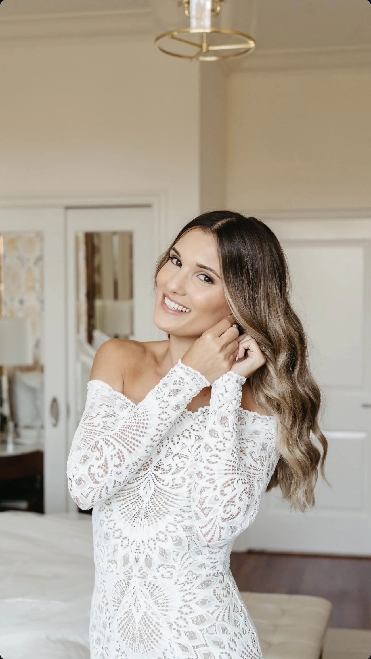 A woman with long wavy hair, smiling, wearing a white lace off-the-shoulder dress, standing indoors in a well-lit room.