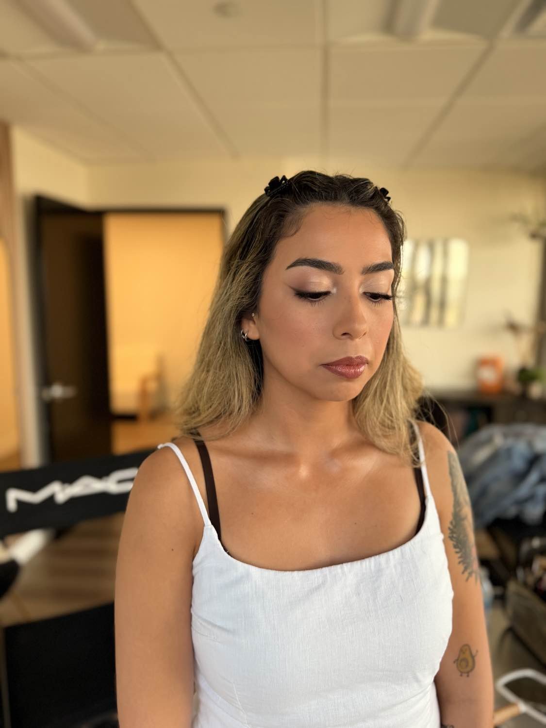 A young woman with shoulder-length blonde hair wearing a white tank top with black straps, makeup, earrings, and tattoos on her arm, looking down with a neutral expression in a room with a blurred background.