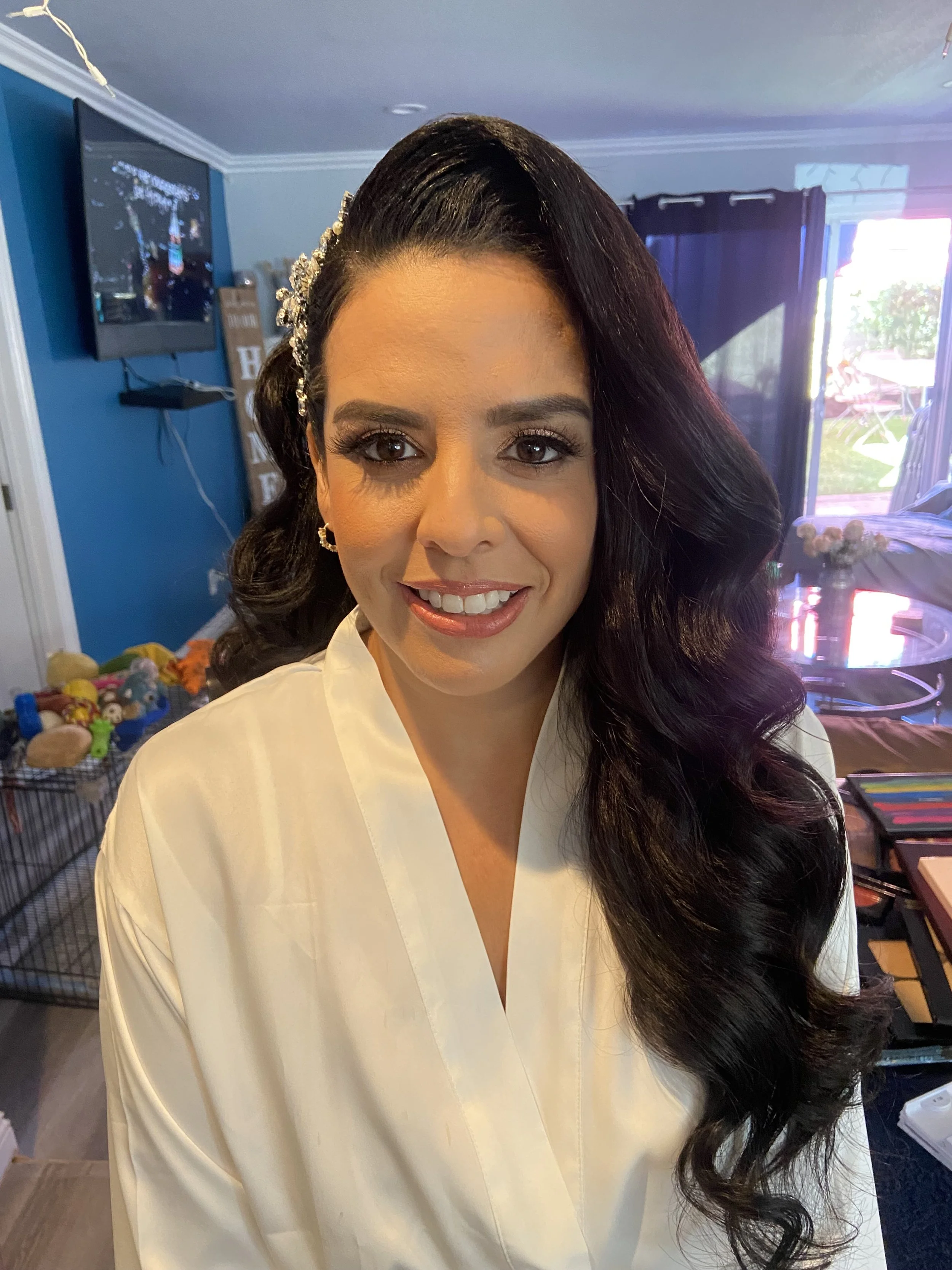 A woman with long, dark hair styled in side curl, wearing a white top, is smiling in a room with a blue wall, TV, and window with dark curtains, while looking at the camera.