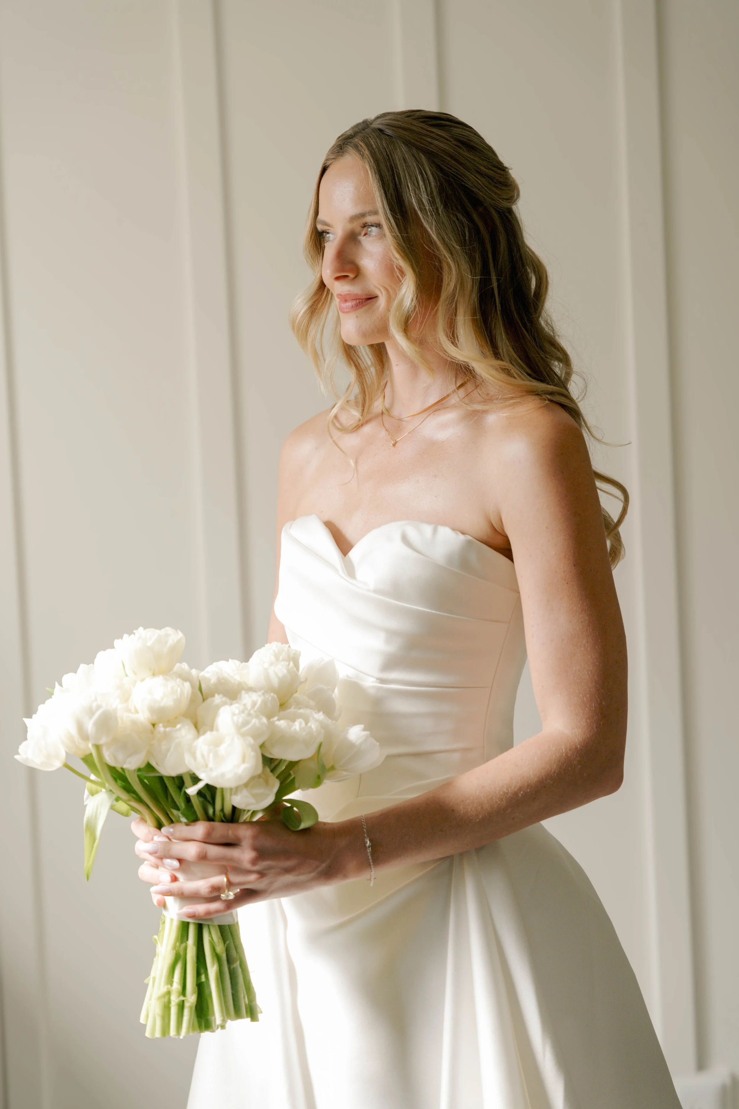 A woman in a strapless white wedding dress holding a bouquet of white flowers, with her hair styled in loose curls, standing indoors in front of a light-colored wall.