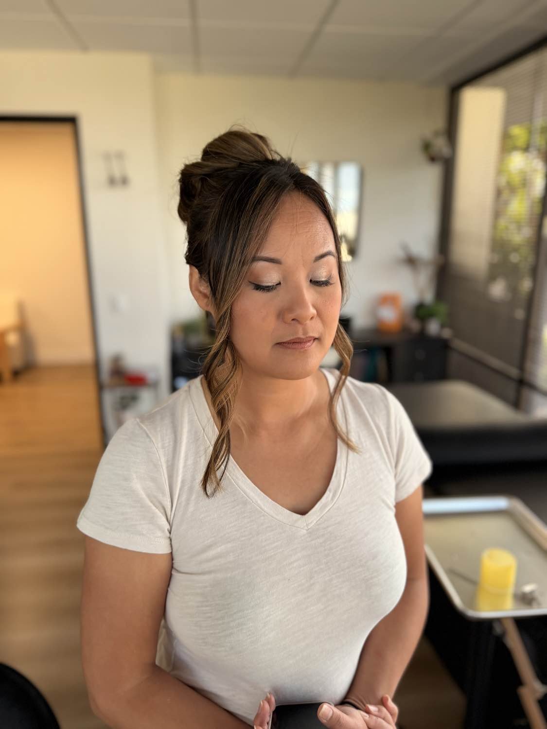 A woman standing indoors with her eyes closed, wearing a white t-shirt, with a mirror and window in the background.