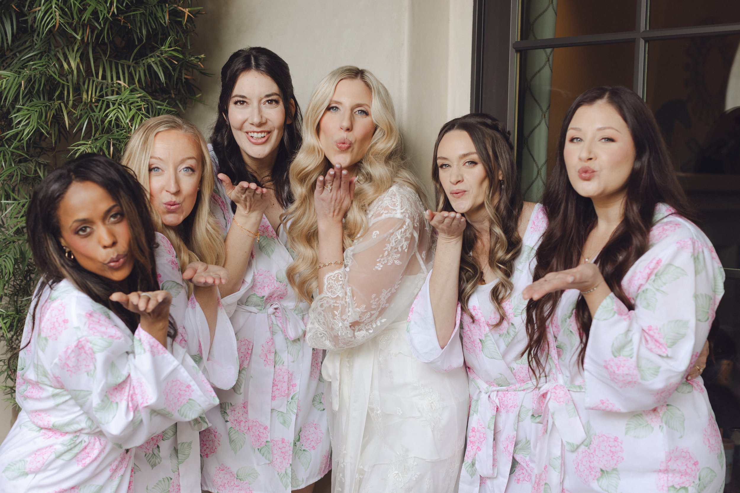 Group of six women, one in wedding dress and the others in matching pajamas, blowing kisses in a celebratory pose.