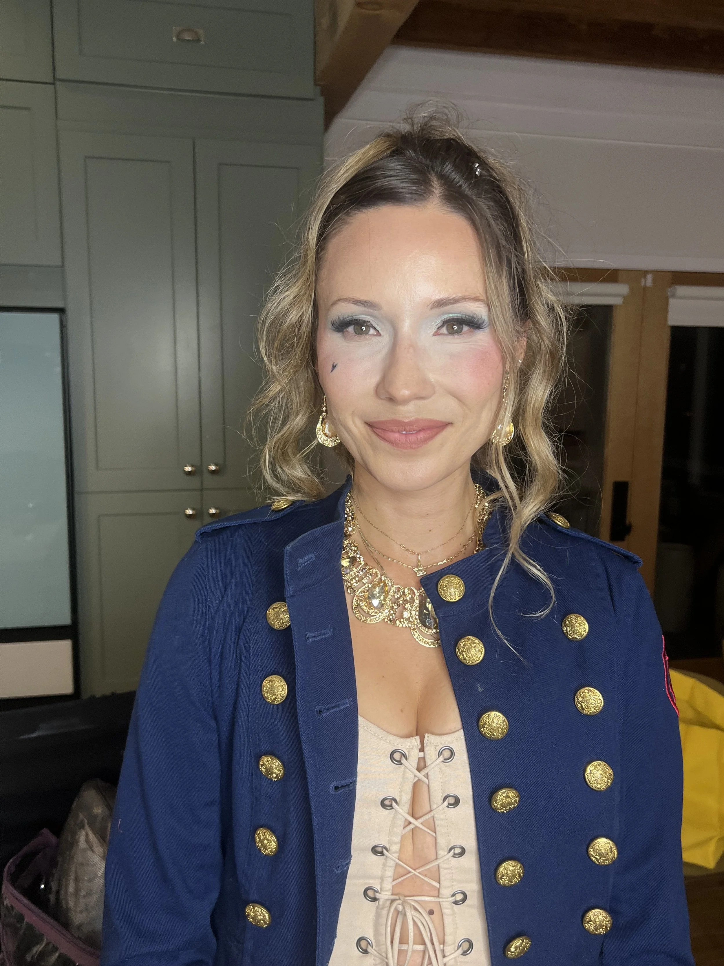 A woman smiling, wearing a navy blue jacket with gold buttons, a beige lace-up top, and gold jewelry, standing in a room with green cabinets and wooden walls.