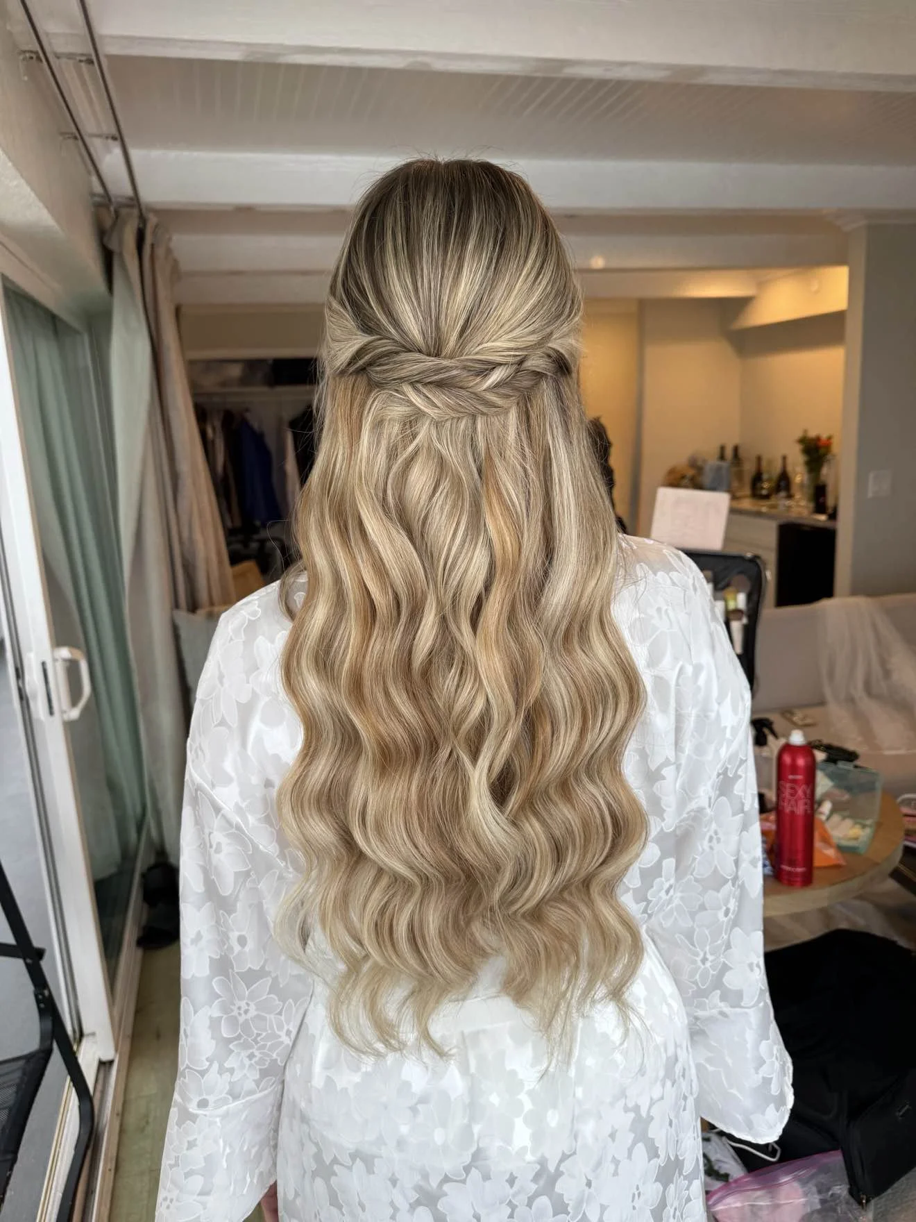 Back view of a woman with long, wavy blonde hair styled in a half-up, twisted braid, wearing a white floral-patterned robe, standing indoors in a room with various items, including a table with bottles and a red spray can.