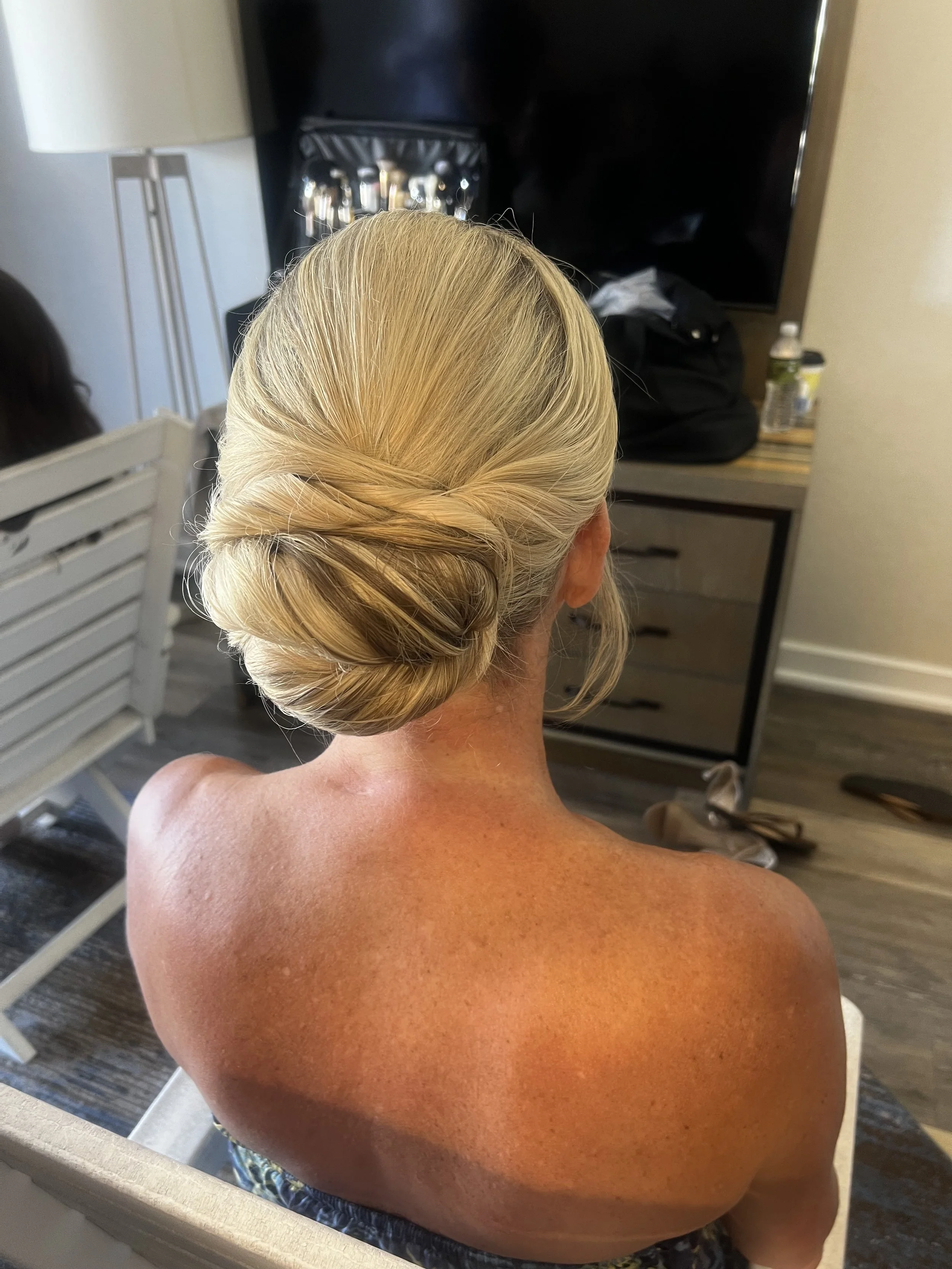 Back of a blonde woman's head with hair styled in an elegant updo, sitting topless in a room with a TV, lamp, and a table with bottles and a black bag.