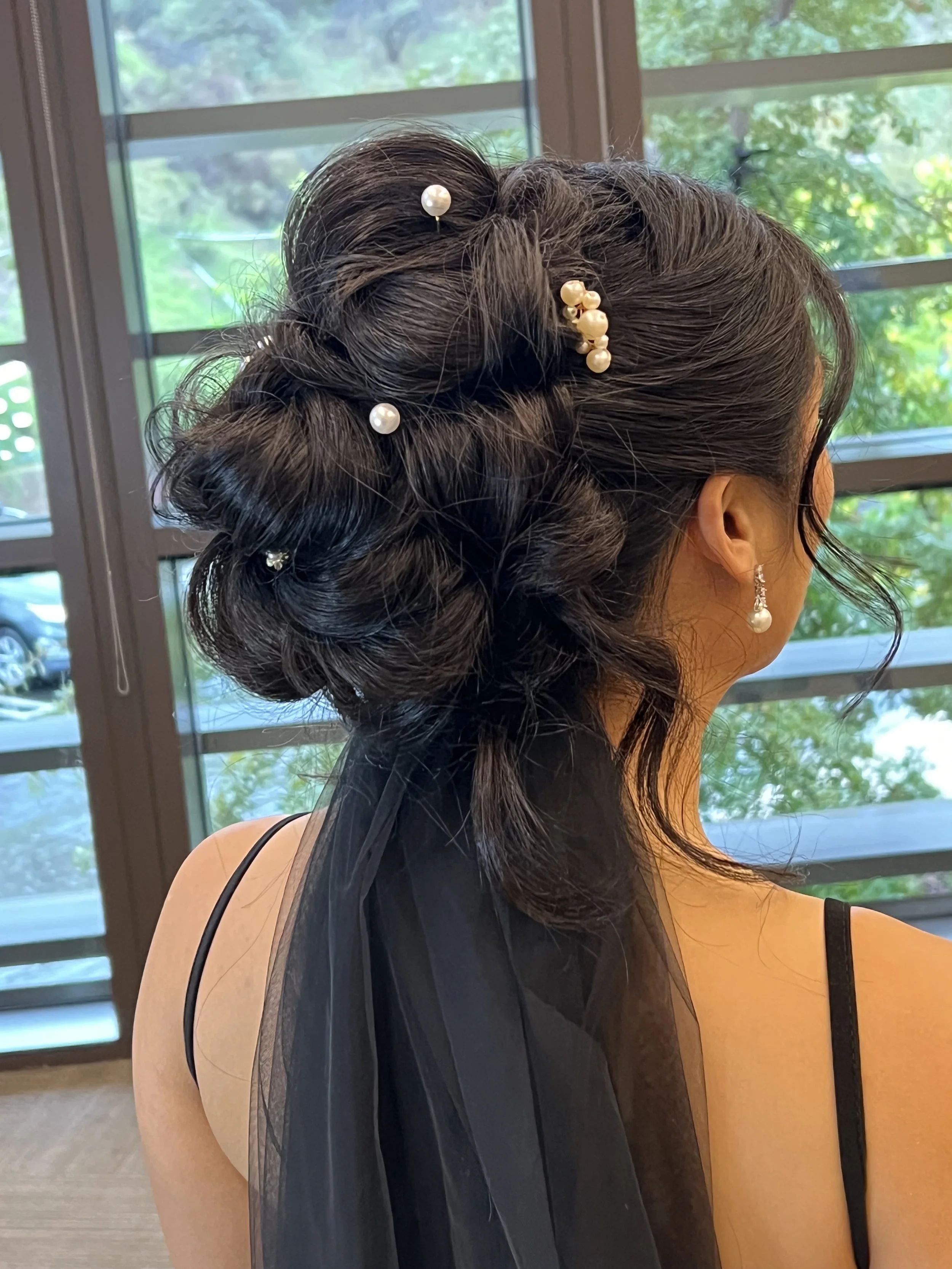 A woman with dark hair styled in an elegant updo, decorated with pearl hairpins and jewelry, looking out a window with greenery outside.