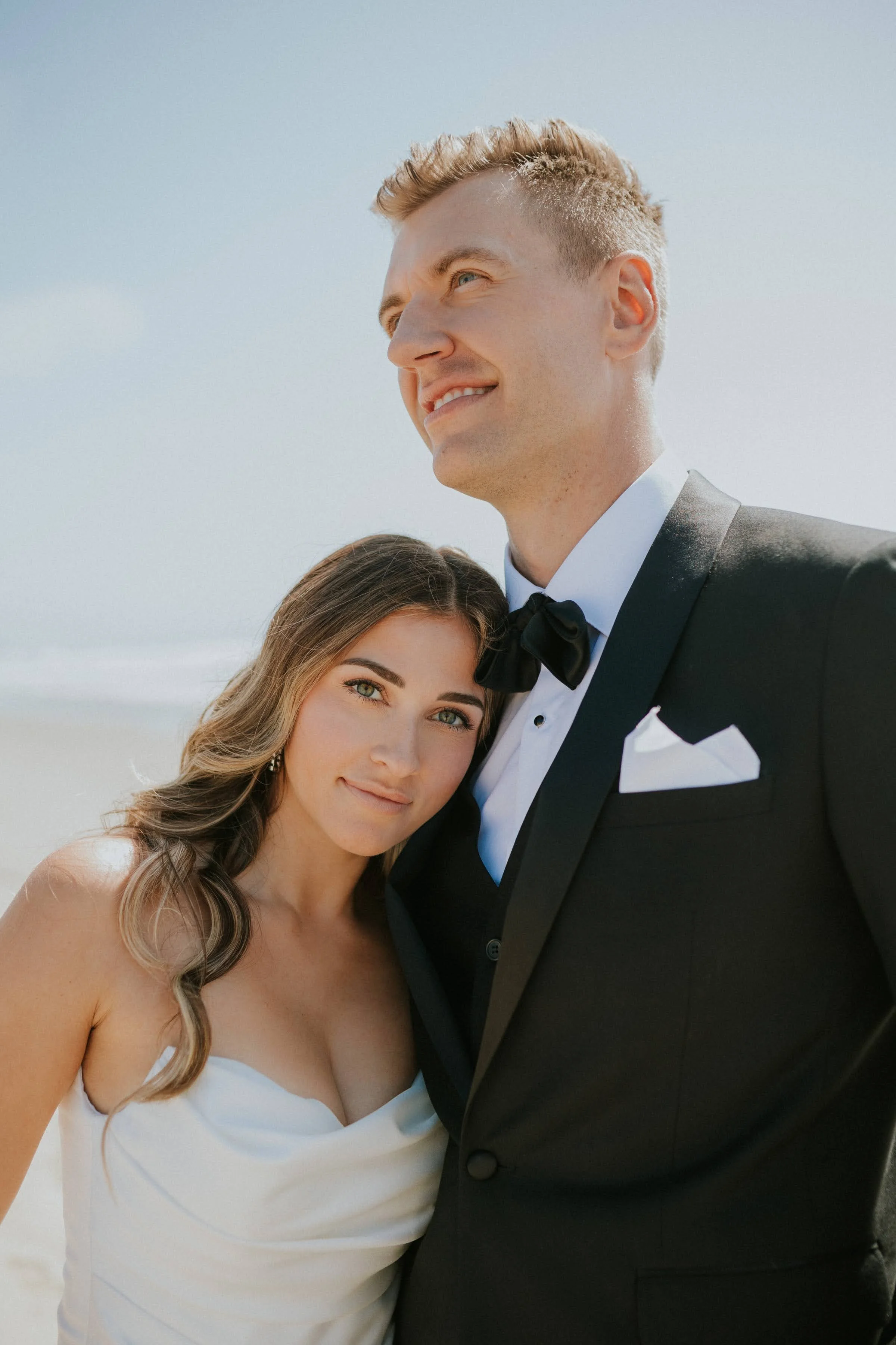 A smiling young woman in a white dress and a young man in a black tuxedo with a bow tie, standing close together outdoors with the sky in the background.