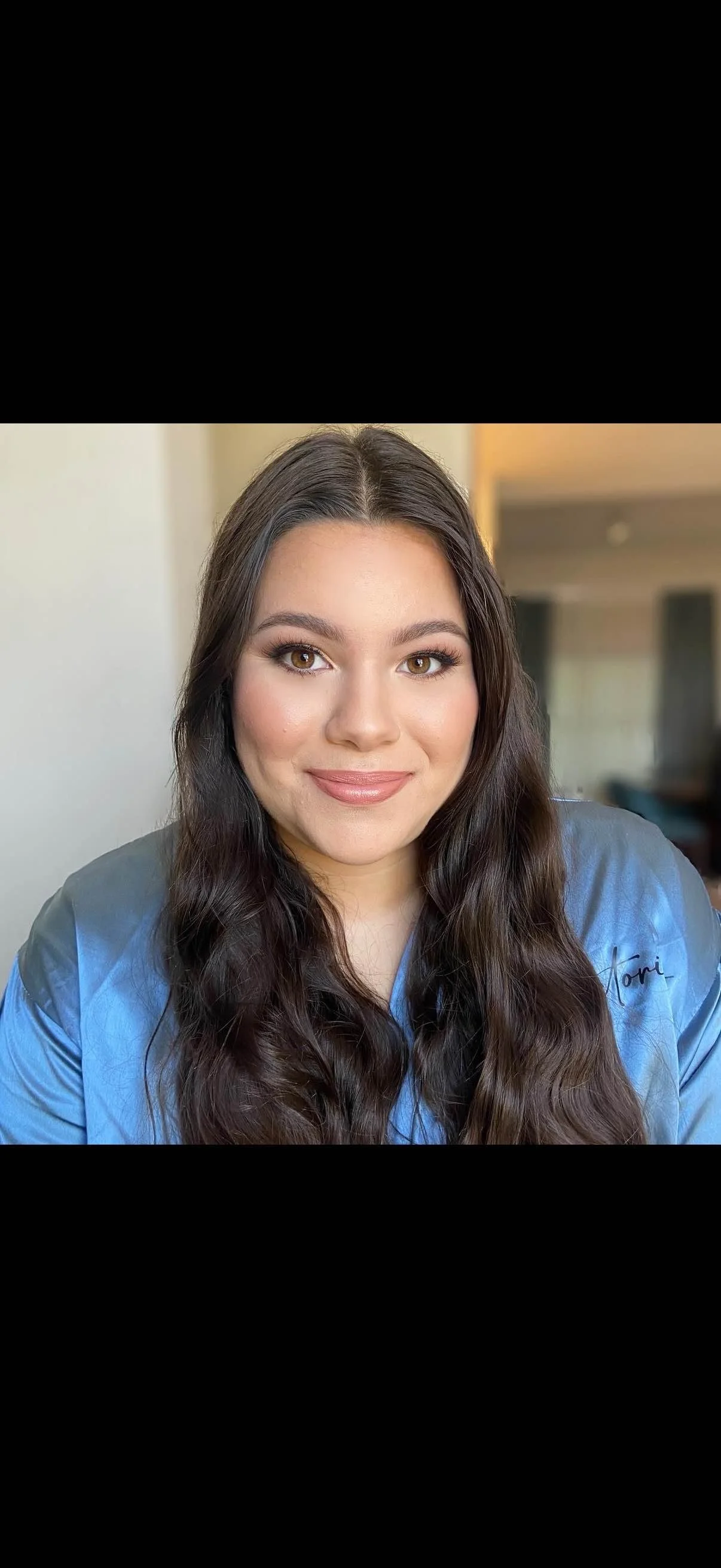 Portrait of a young woman with long dark wavy hair, brown eyes, and a light pink lipstick, wearing a blue shirt with the name "Tori" embroidered on it, smiling indoors.