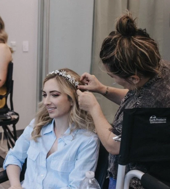 A woman in a dark shirt styles a young woman’s blonde hair and floral headband, who is sitting and smiling.