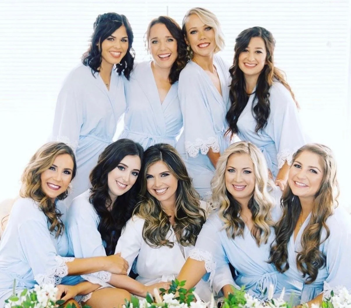 Group of nine women dressed in matching light blue robes, smiling and sitting together near white flowers, in a bright room with a window background.