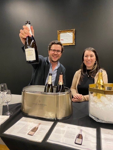 A man and a woman standing behind a bar with Champagne bottles on ice. Man is holidng a bottle of Louis Roederer Starck Champagne.