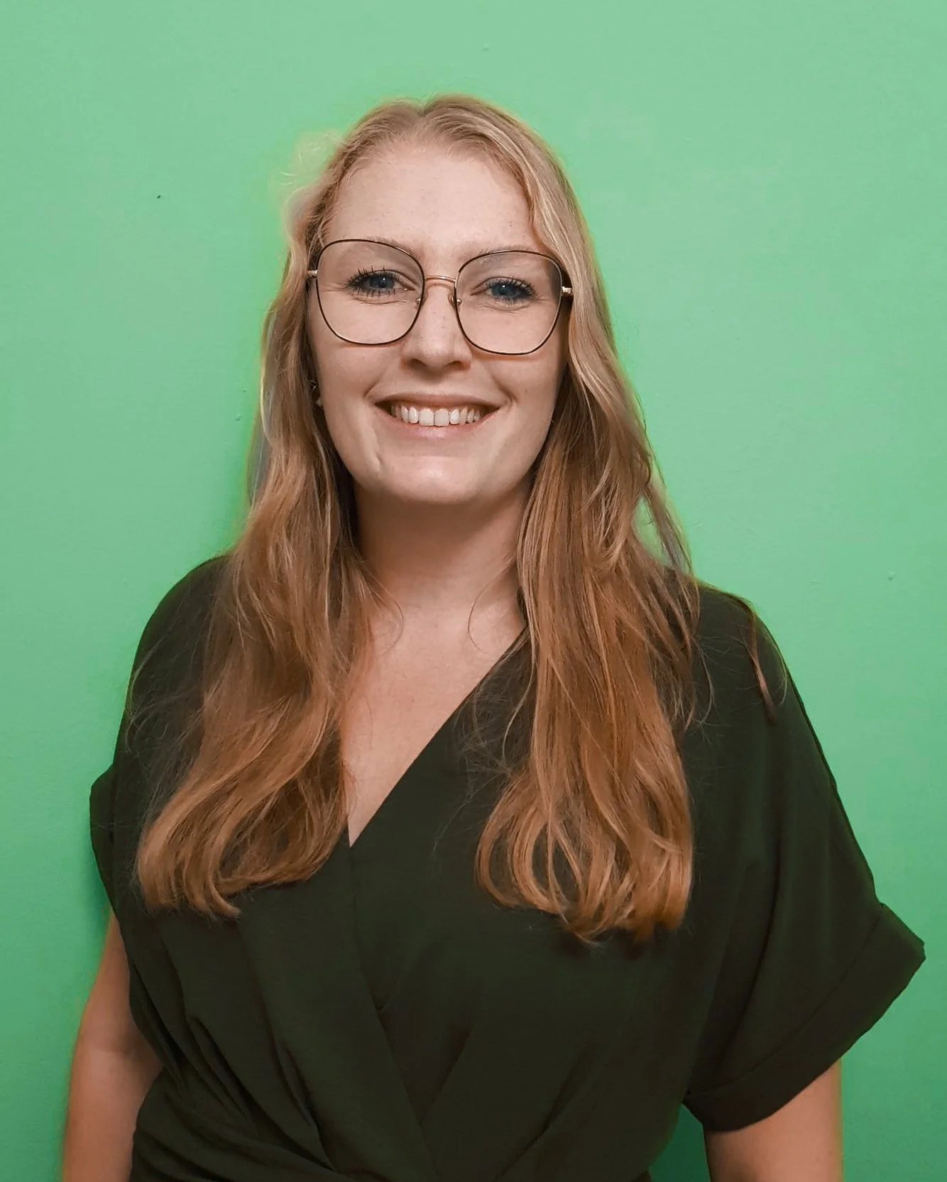 A woman with long, light brown hair and glasses, smiling, wearing a black top, standing against a purple background.