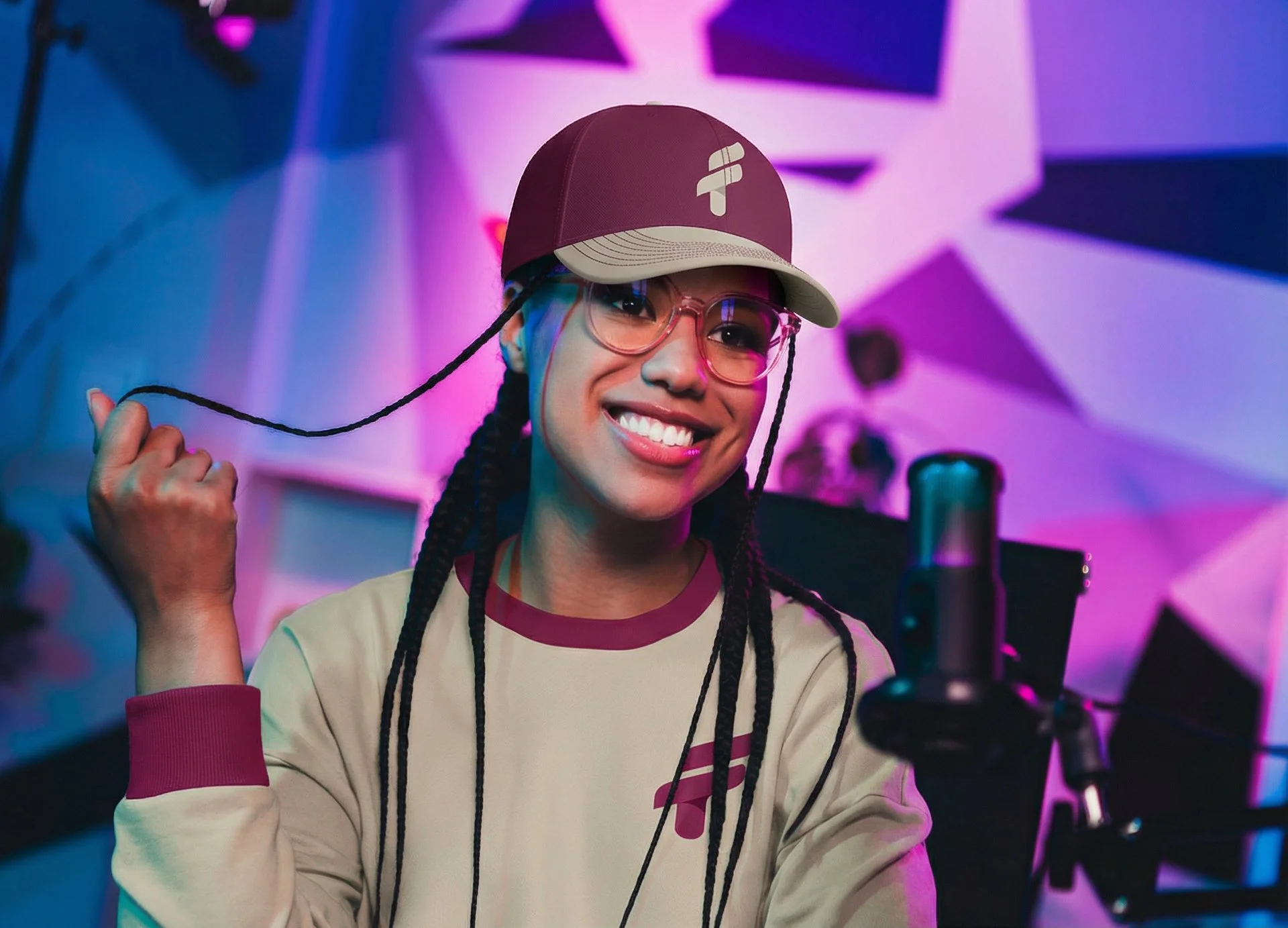 A smiling young woman with braided hair, wearing a beige baseball cap, pink glasses, and a beige sweatshirt with red accents, in a colorful, neon-lit recording studio.