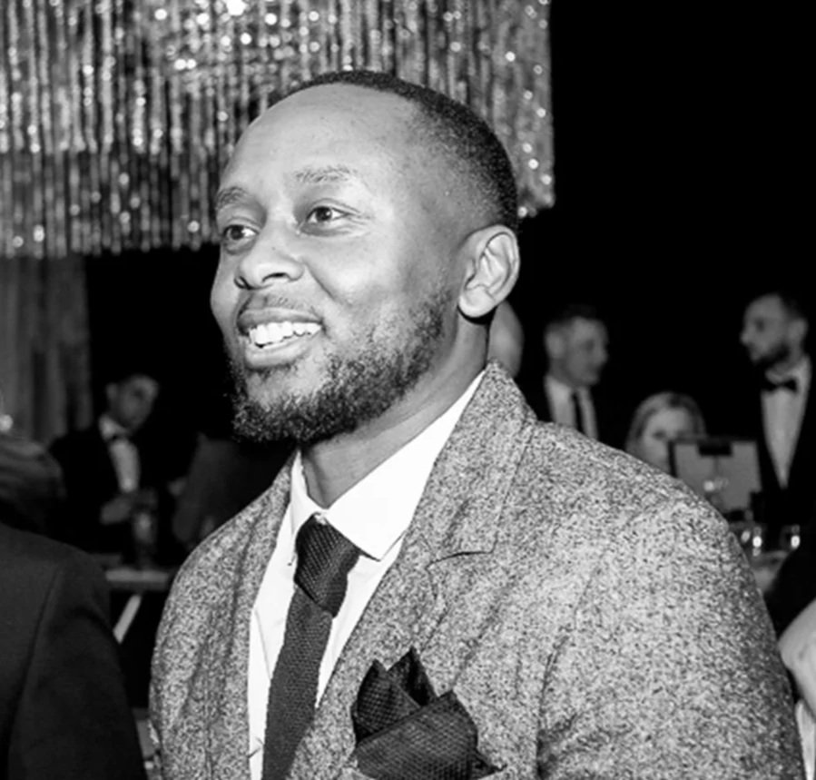 A smiling man in a suit and tie at a formal event, with people in the background, some in tuxedos, and shiny decorations overhead.