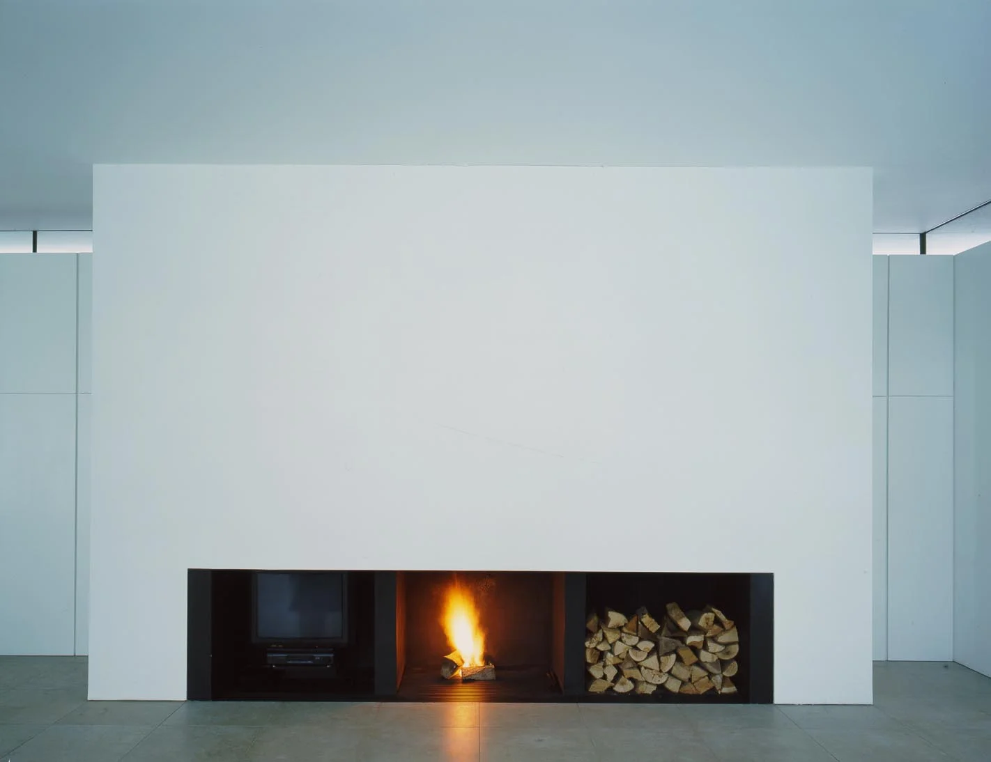The concrete structural chimney breast is a clear and pure presence in the house . The wide horizontal opening includes a TV , fireplace and a log store. 