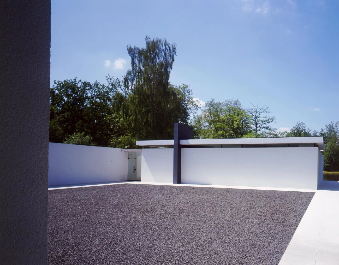 The west side of the courtyard where the main entrance is located between the living room and bedroom wings . Here one sees the floating roof plane above the clerestory and the vertical grey chimney element that contains the flue and drainage pipework. 