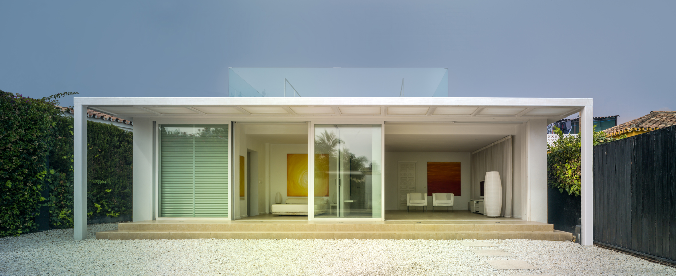 The facade with the glass sliding doors in the central position.All the panels can be stacked in front of the louvers to the left to fully open up the interior to the outside. There is a roof terrace with a blue tinted glass balustrade. 