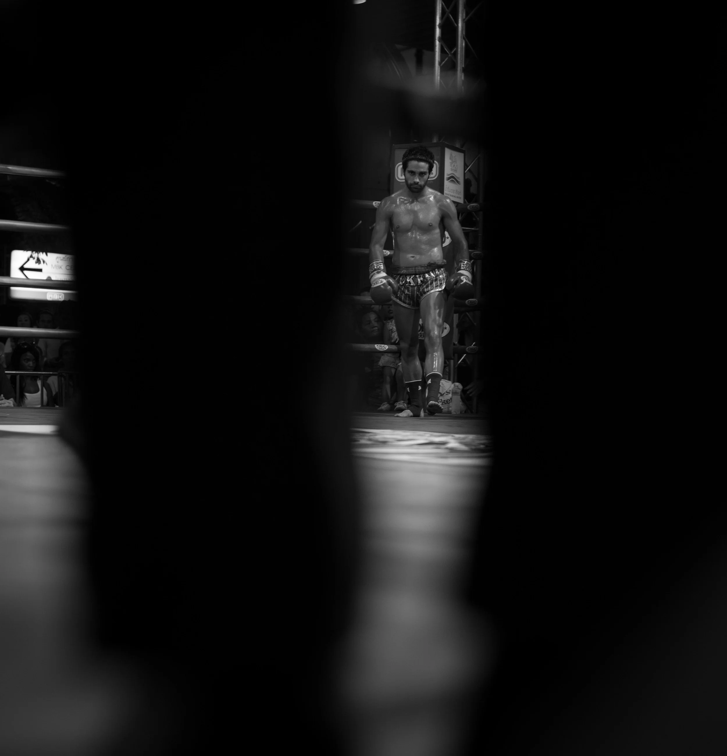 Black and white photo of a boxer with gloves and boxing shorts, standing in a ring made of wood, with audience members in the background. The photo is taken through a small opening or gap.