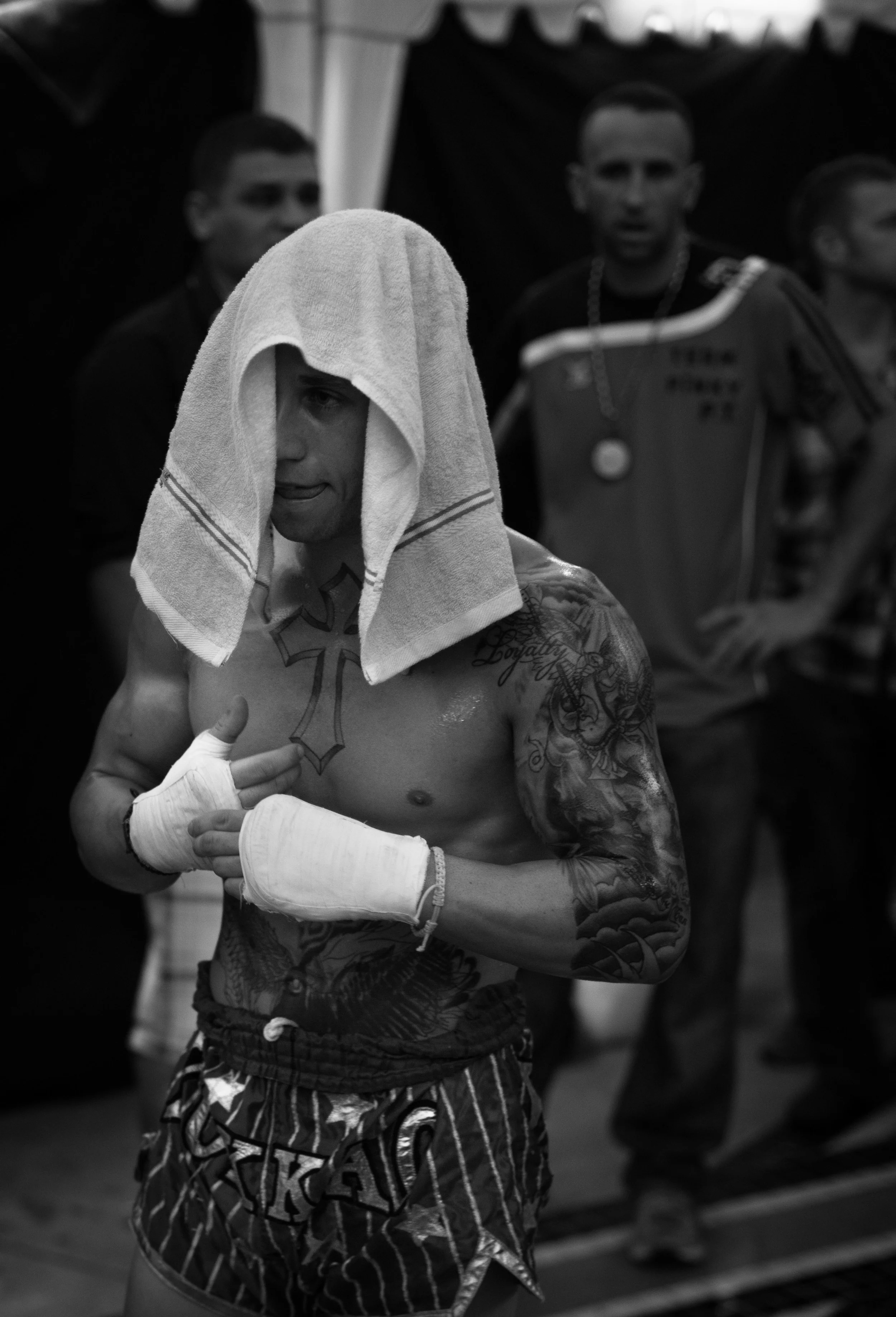 A male boxer with a towel on his head, tattooed upper body, and wearing boxing gloves and shorts, standing in a gym with other people in the background.
