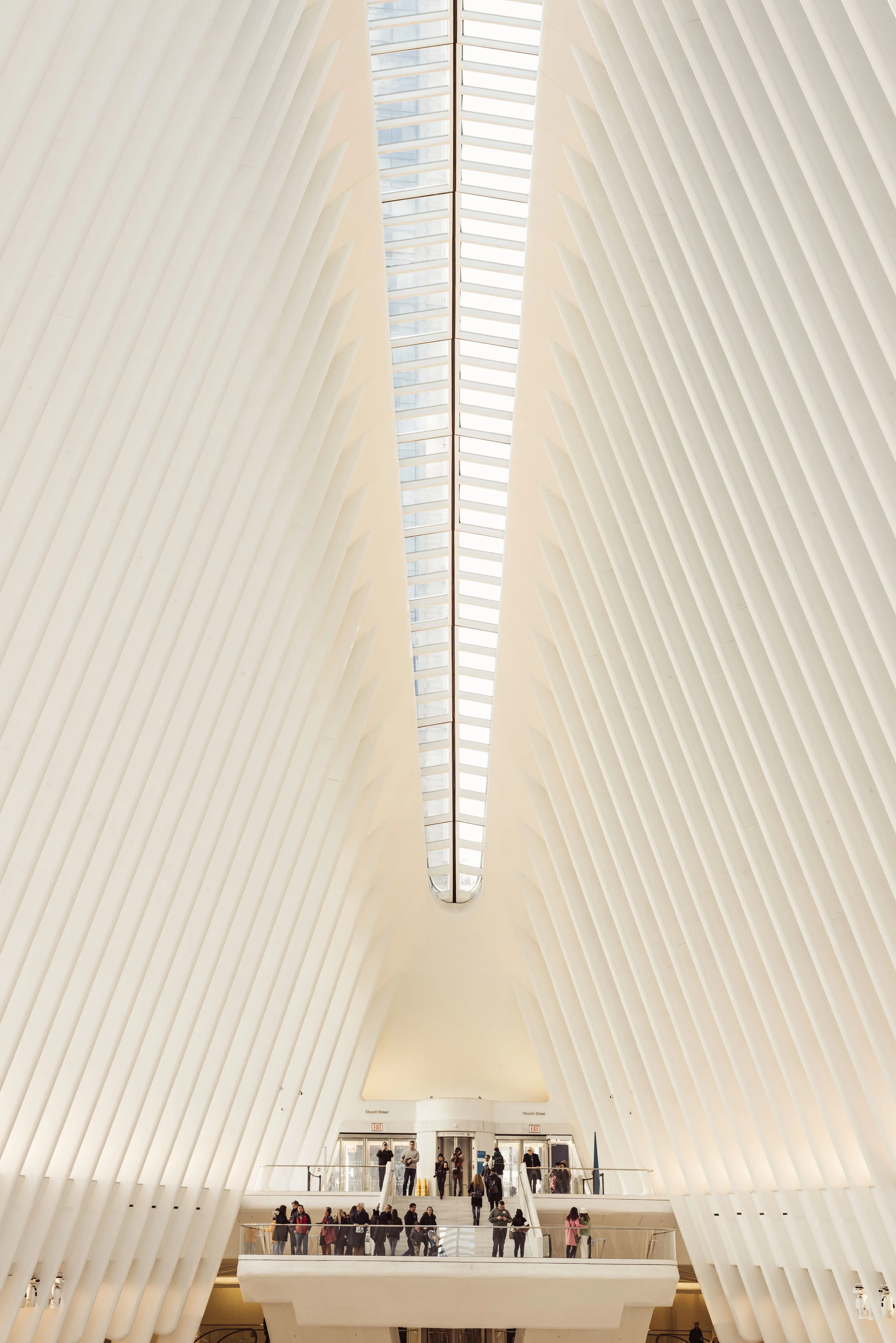 Inside the Oculus, a large architectural structure with white rib-like beams curving along the walls and ceiling. There is an upper level with people walking and gathering near a glass railing, and a lower level with more people, escalators, and visi