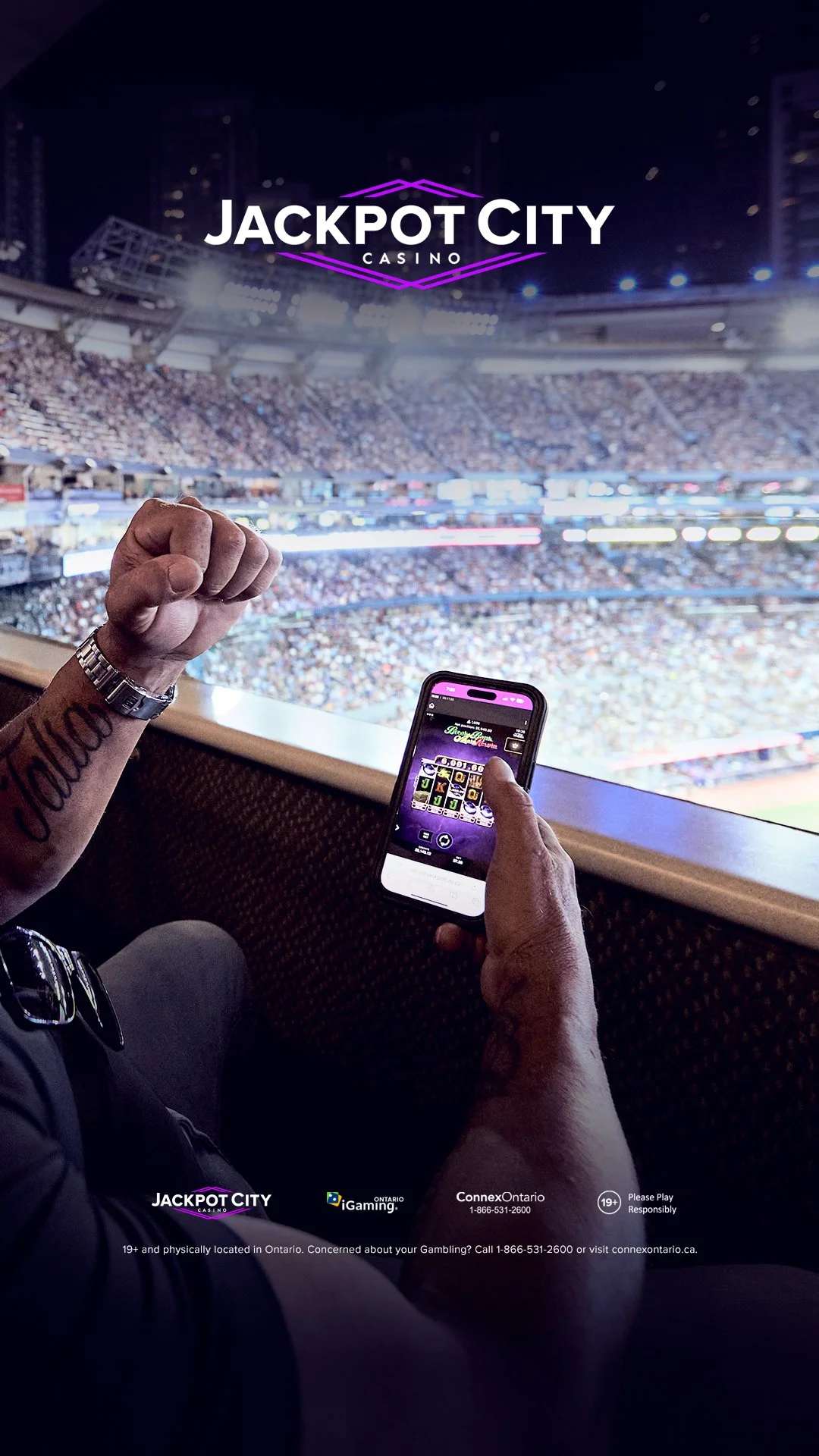 A person holding a smartphone with a slot machine game on the screen, a tattoo on their arm, and a wristwatch, sitting in a stadium filled with spectators during a baseball game, with the JAckpot City Casino logo overlayed at the top.